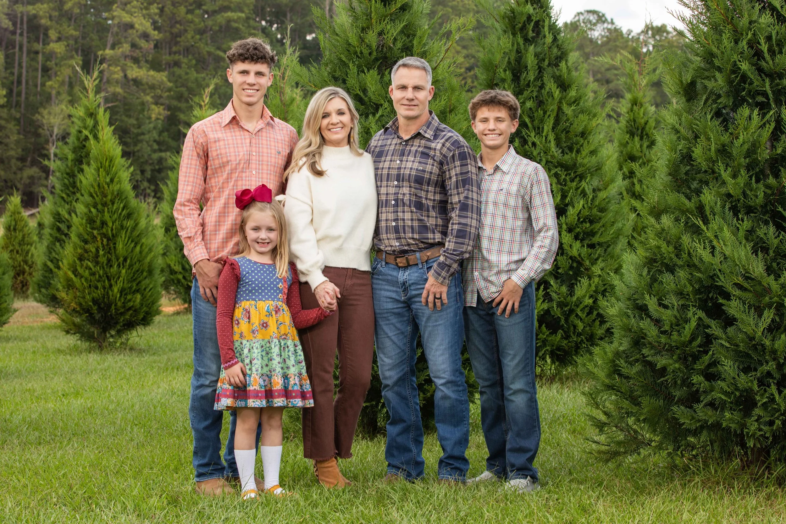 Family of six standing outdoors among green trees, holding hands and smiling at camera.