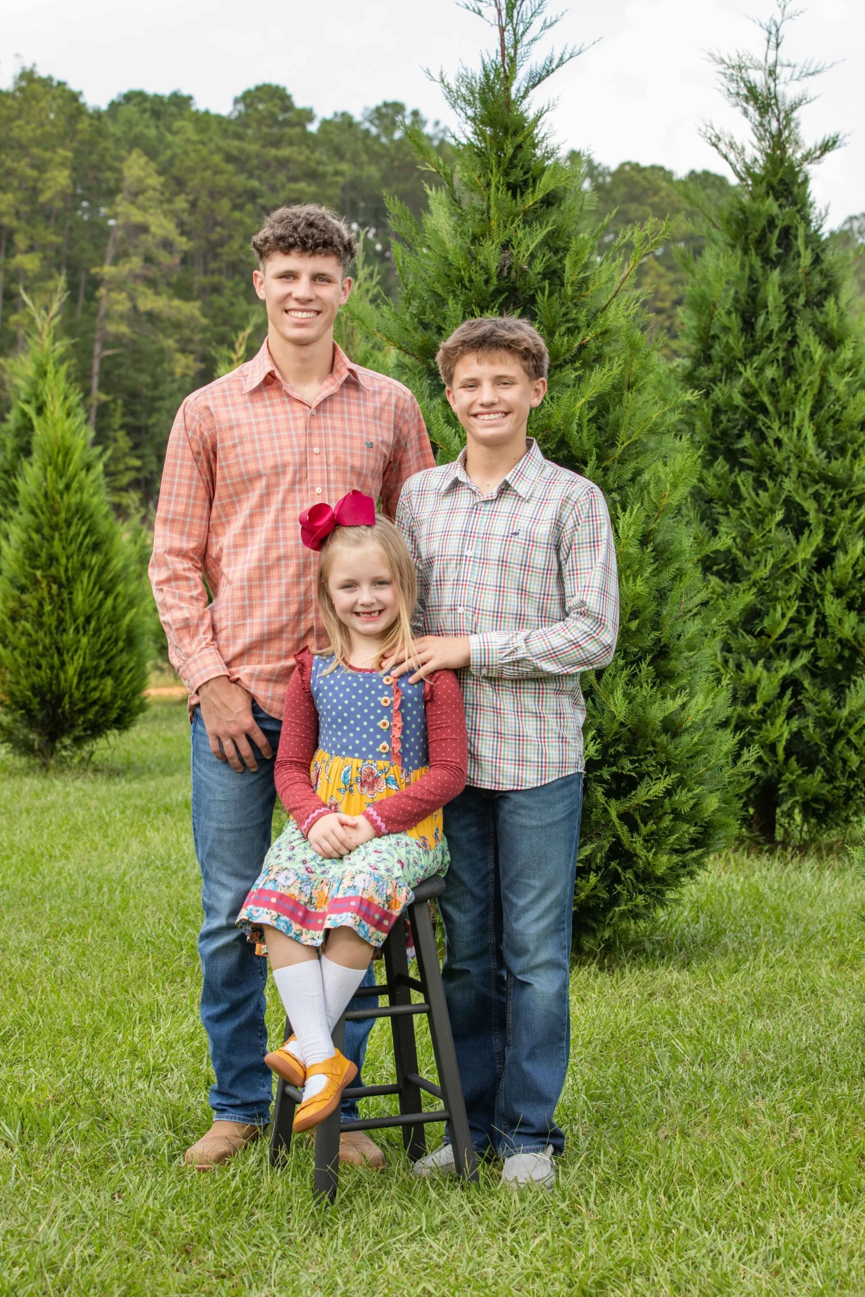 Three children, two boys and a girl, standing outside in a grassy area with trees in the background. The girl is sitting on a small black stool, wearing a colorful dress with a large pink bow in her hair. The boys are standing behind her, smiling at the camera.