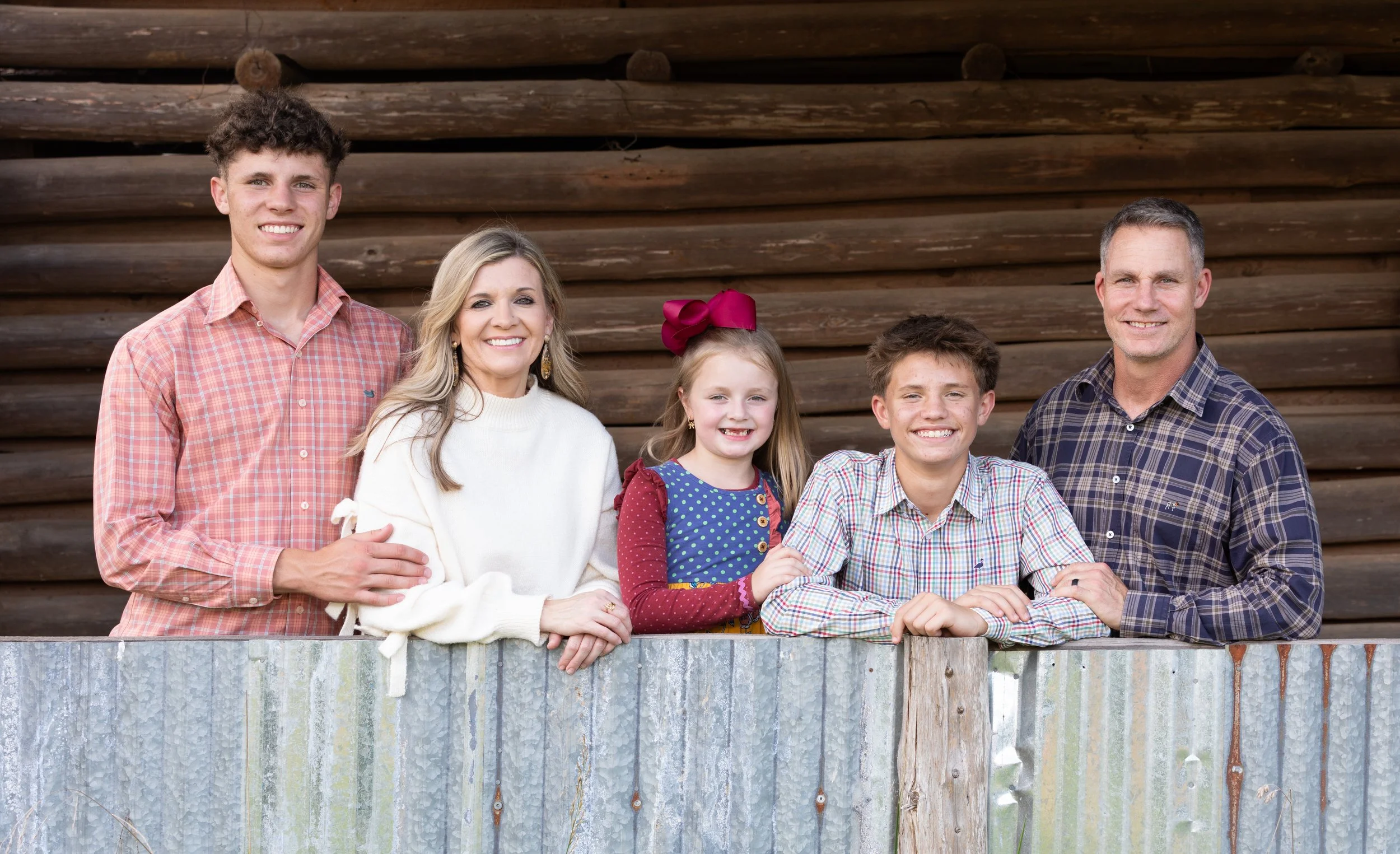 Family of six posing outdoors in front of a wooden building, leaning on a corrugated metal fence.