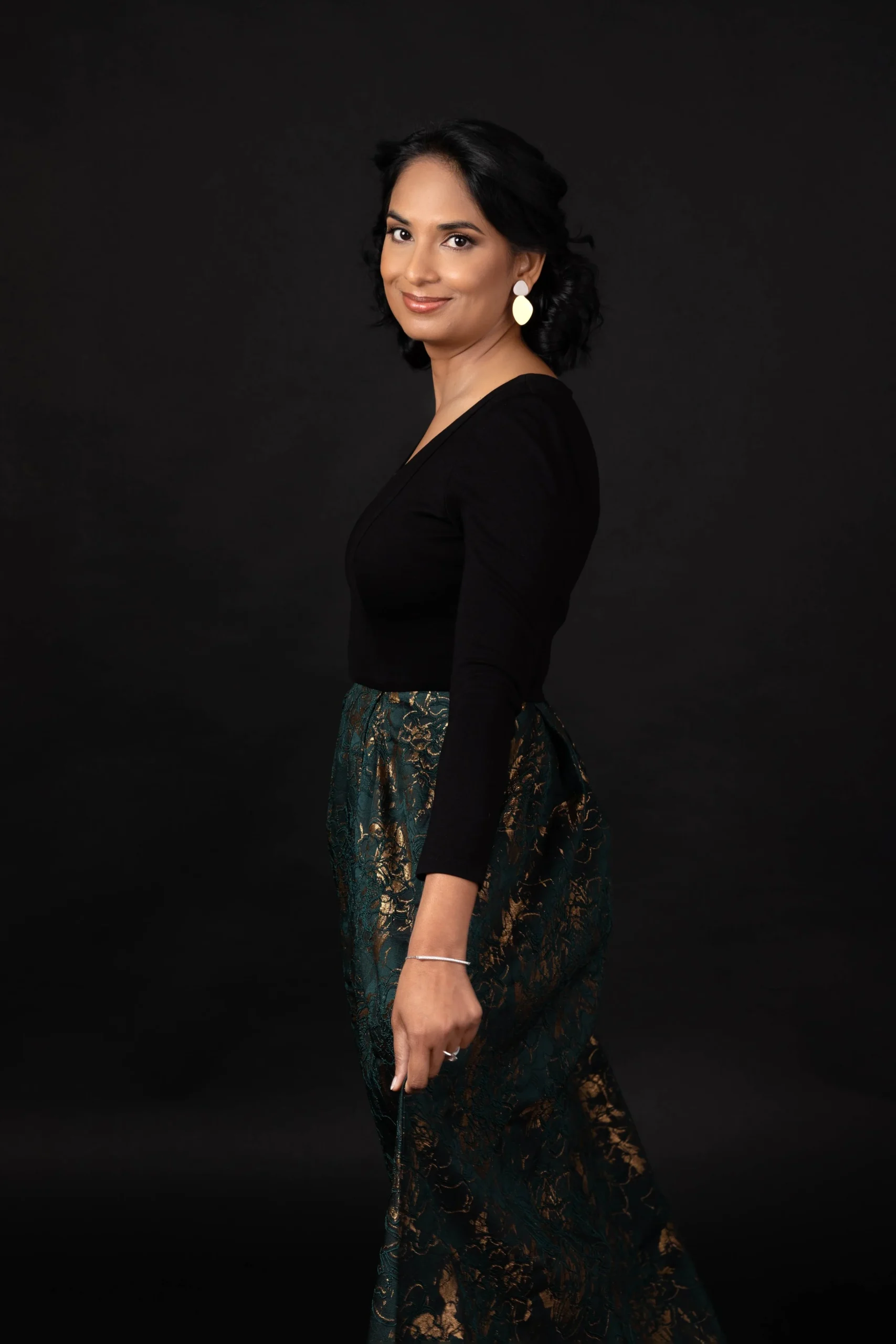 A woman with dark hair and earrings wearing a black top and patterned pants standing against a black background.