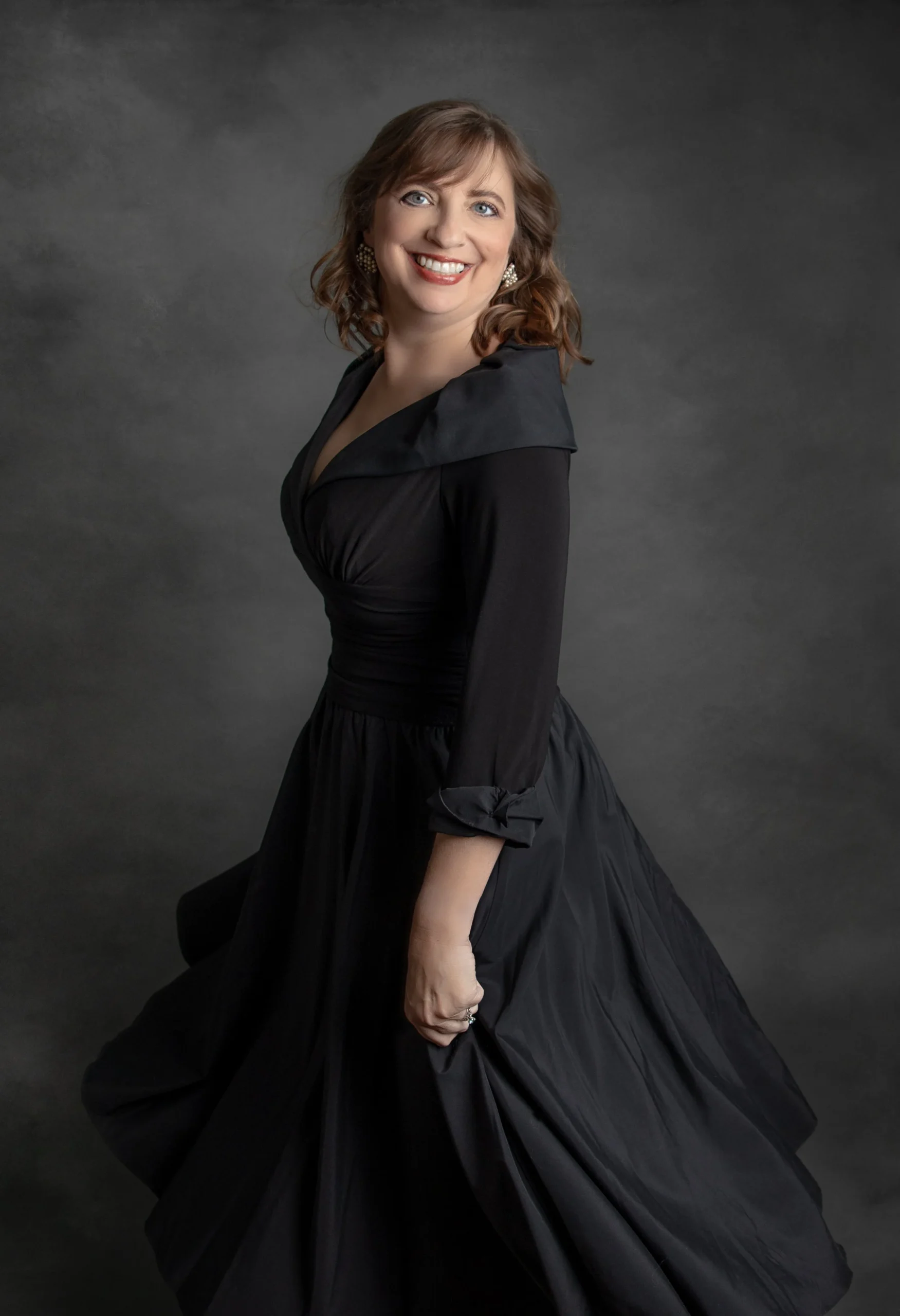 A woman with curly brown hair and blue eyes wearing a long black dress with a wide collar, standing against a dark gray background.