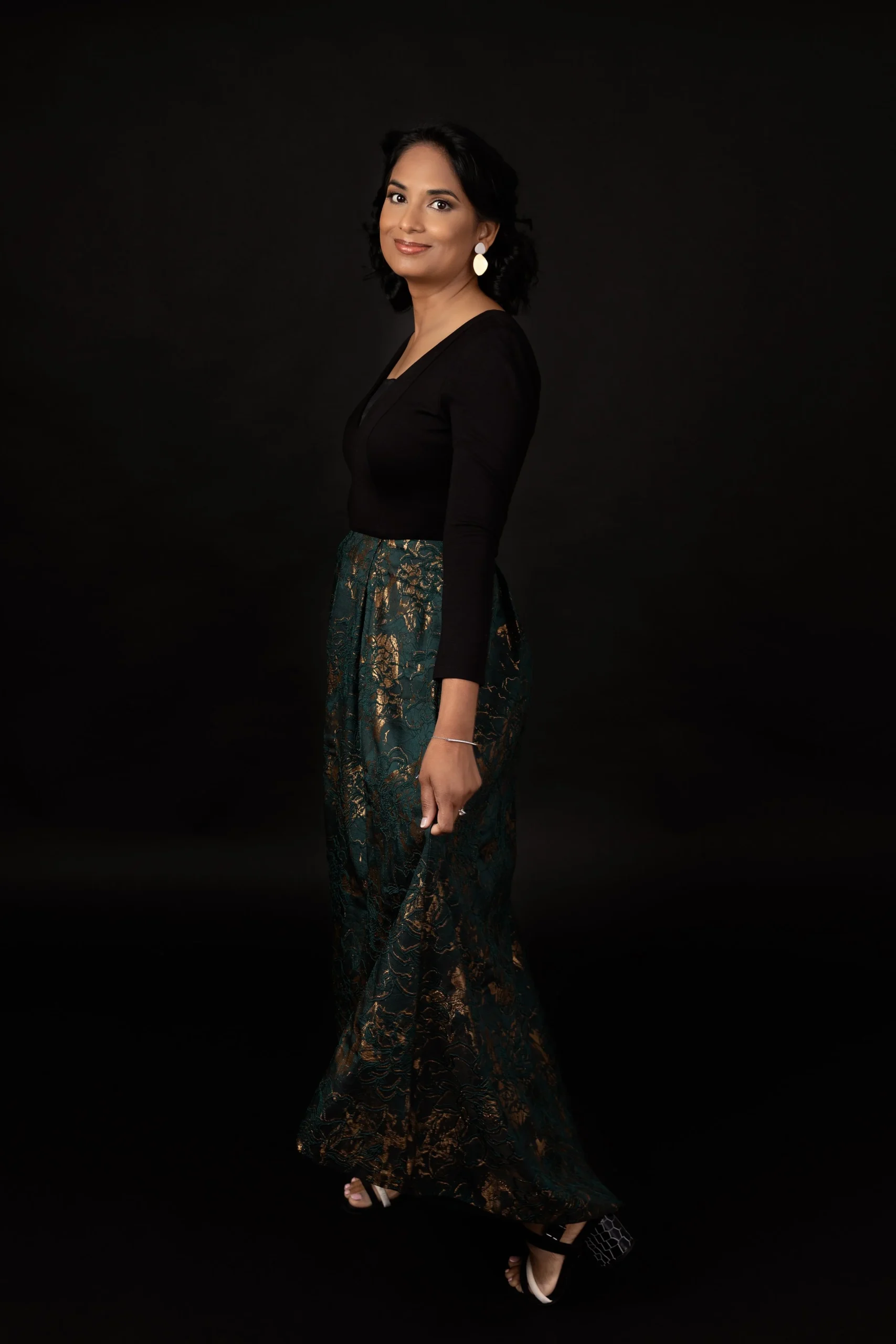 A woman with dark hair and earrings wearing a black top and patterned green and gold wide-leg pants standing against a dark background.