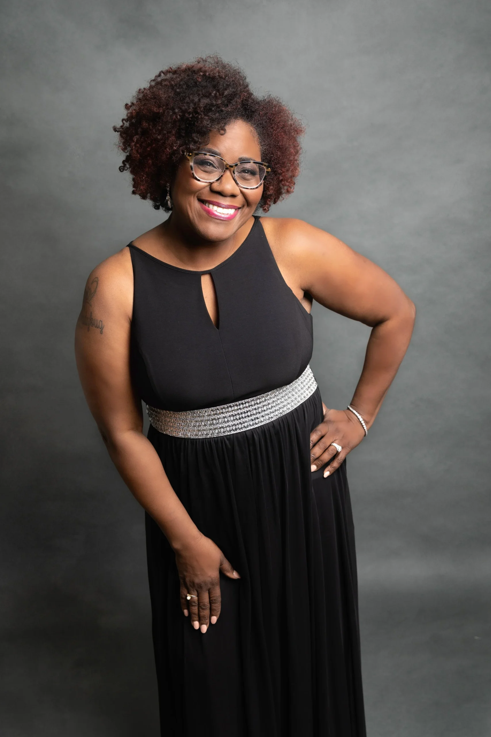A smiling African American woman in a black dress with a beaded waist, standing against a gray background, with glasses, short curly hair, and jewelry.