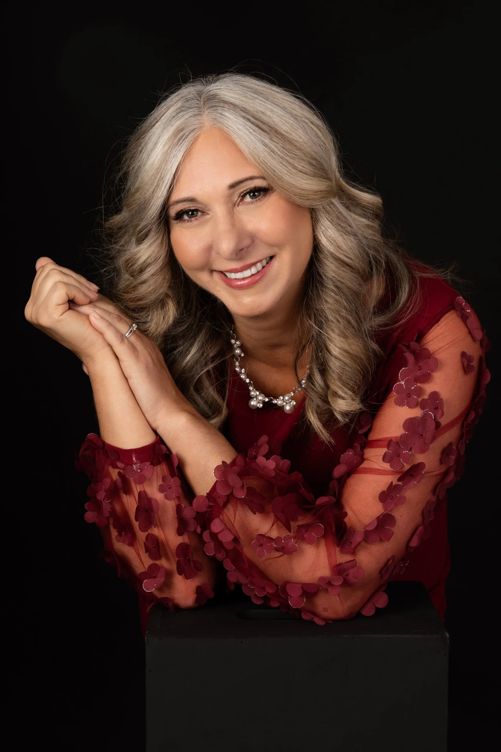 A smiling woman with long, curly gray hair, wearing a red dress with sheer sleeves decorated with red flowers, and a pearl necklace, posing against a black background.