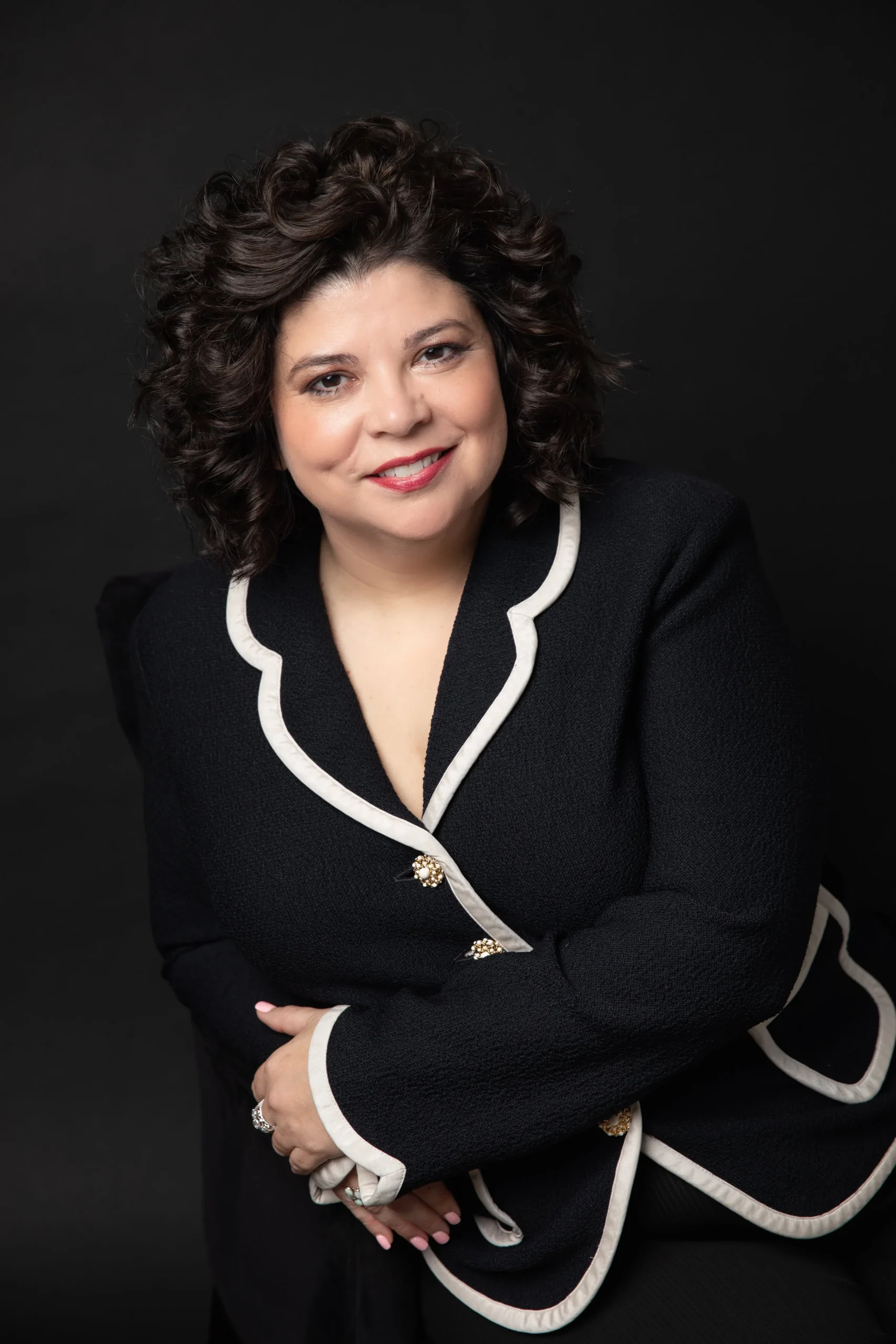 A woman with curly dark brown hair, wearing a black blazer with white trim and decorative buttons, is smiling and sitting against a dark background.