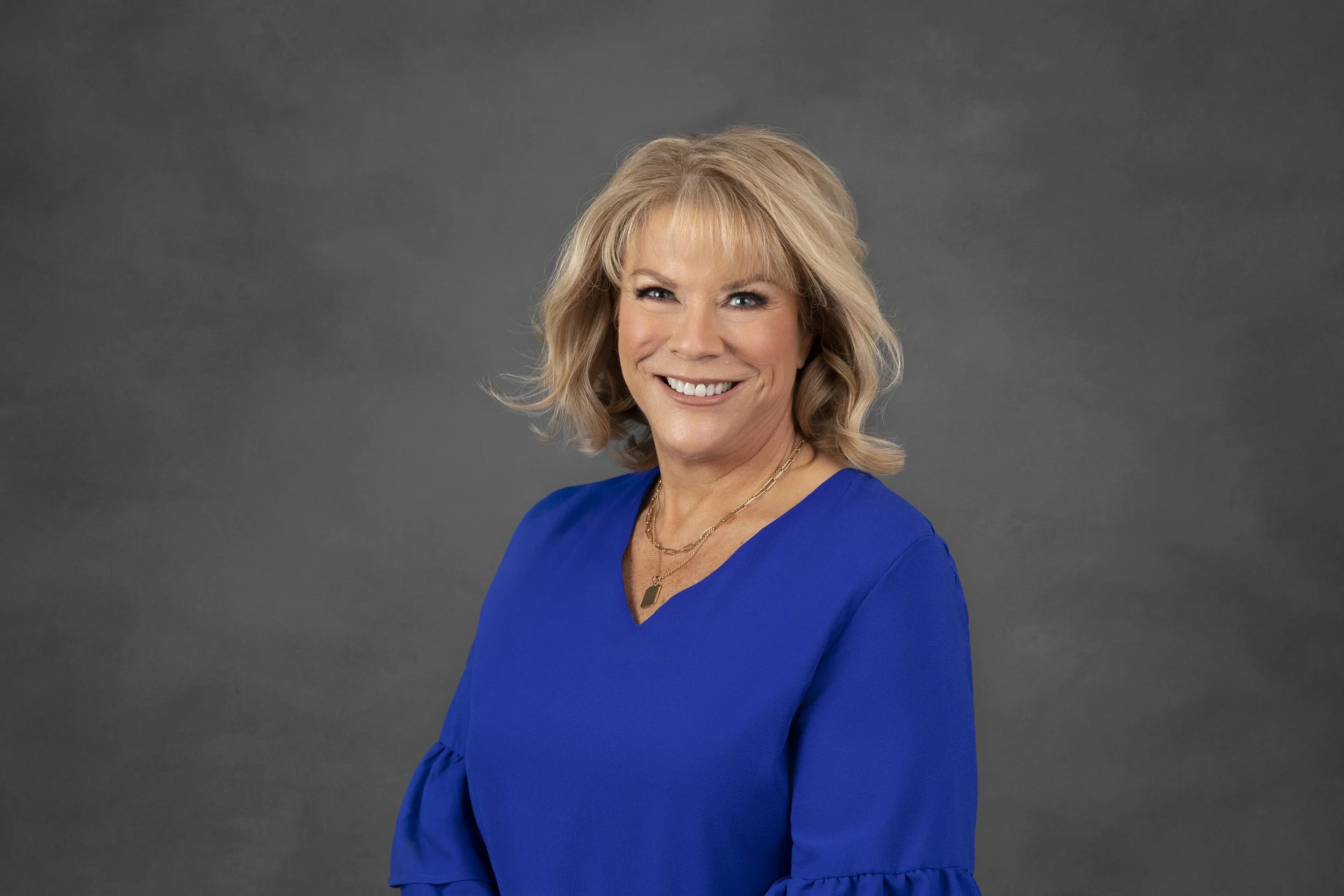 Portrait of a woman with blonde hair wearing a blue top, winking and smiling against a gray background.