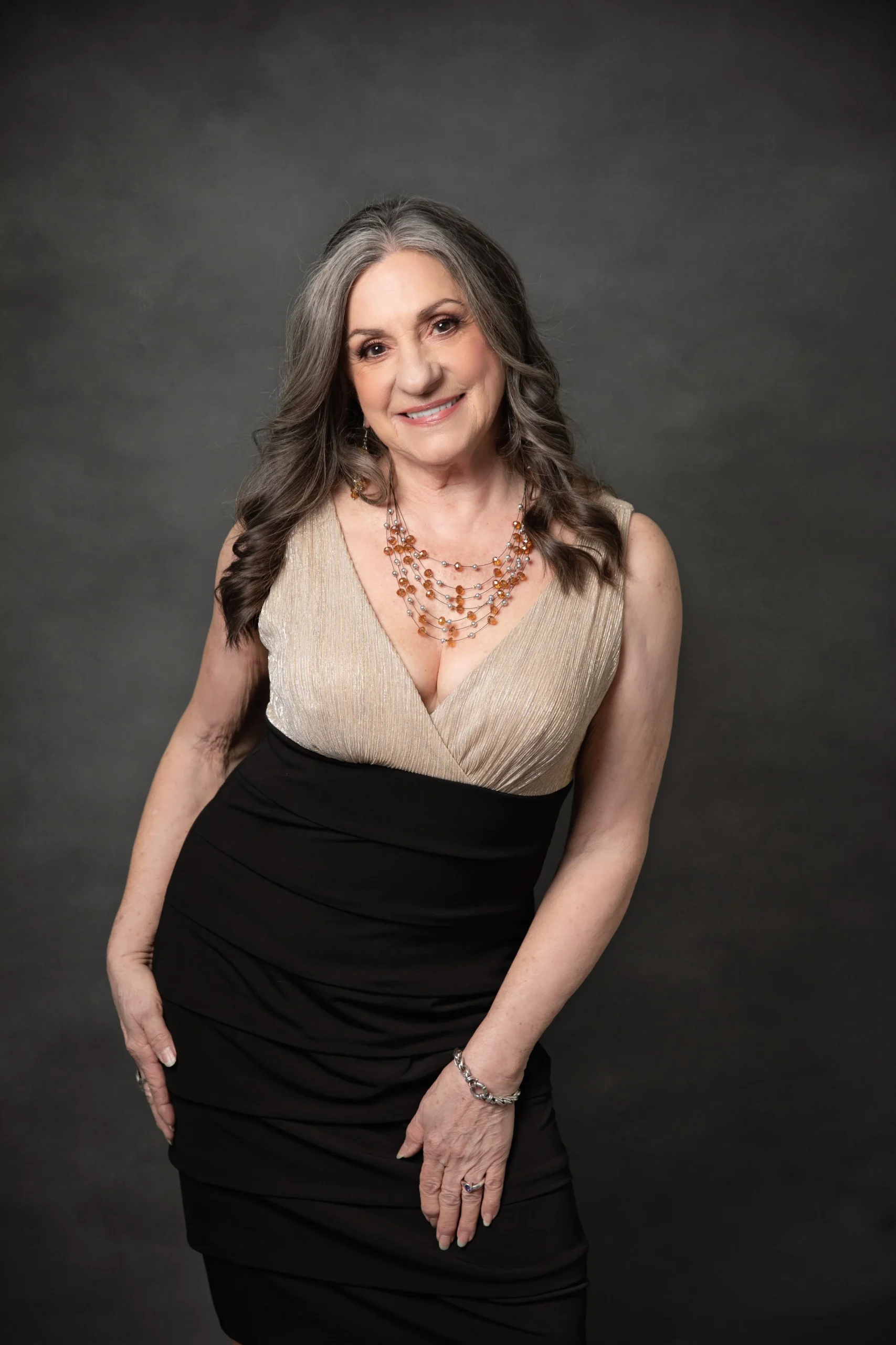 An elderly woman with gray hair styled in loose waves, wearing a sleeveless gold and black dress with layered necklaces and a bracelet, smiling softly against a dark gray background.