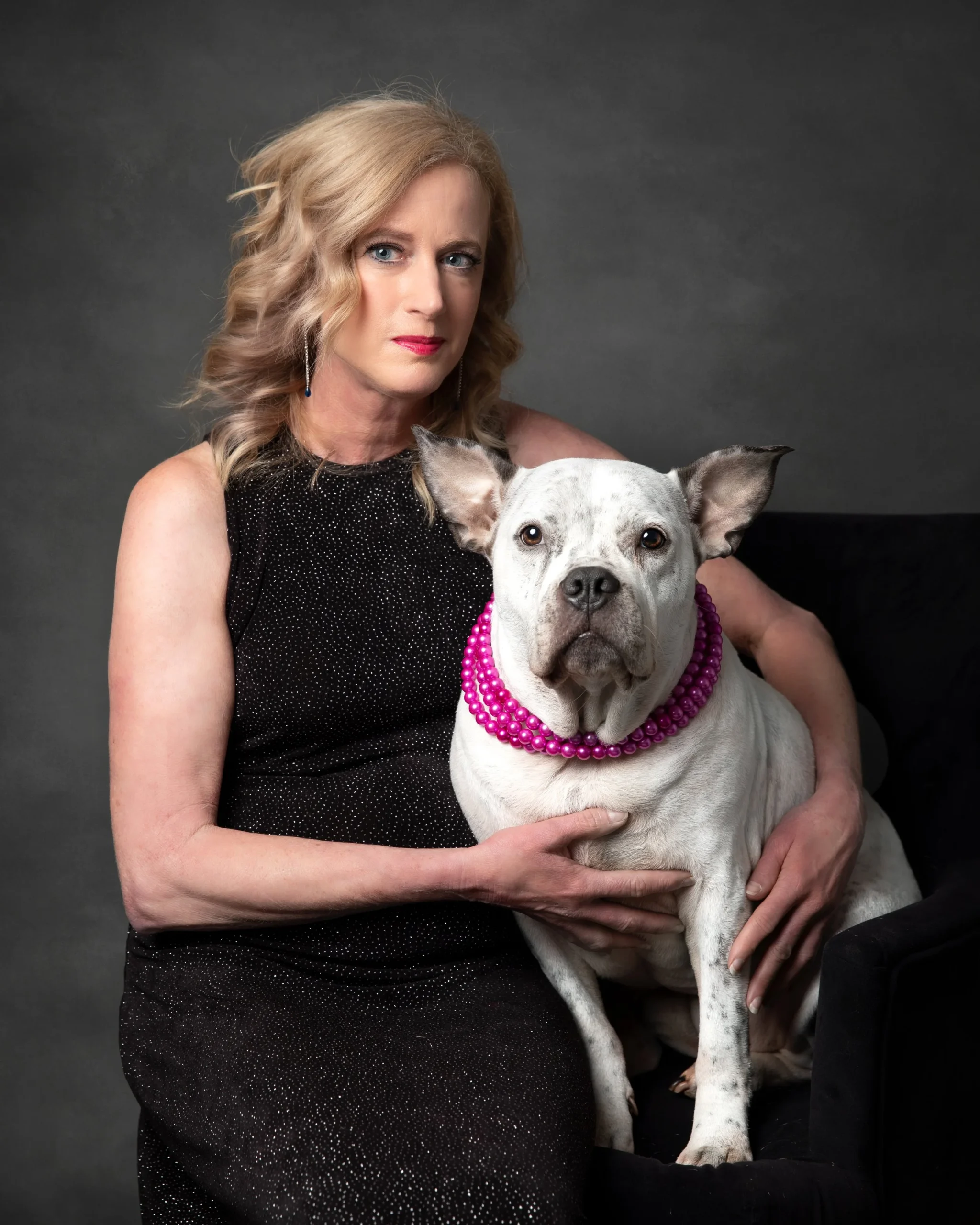 A woman with blonde hair in a black dress sitting next to a white dog wearing a pink beaded necklace and a pink beaded collar, against a gray background.