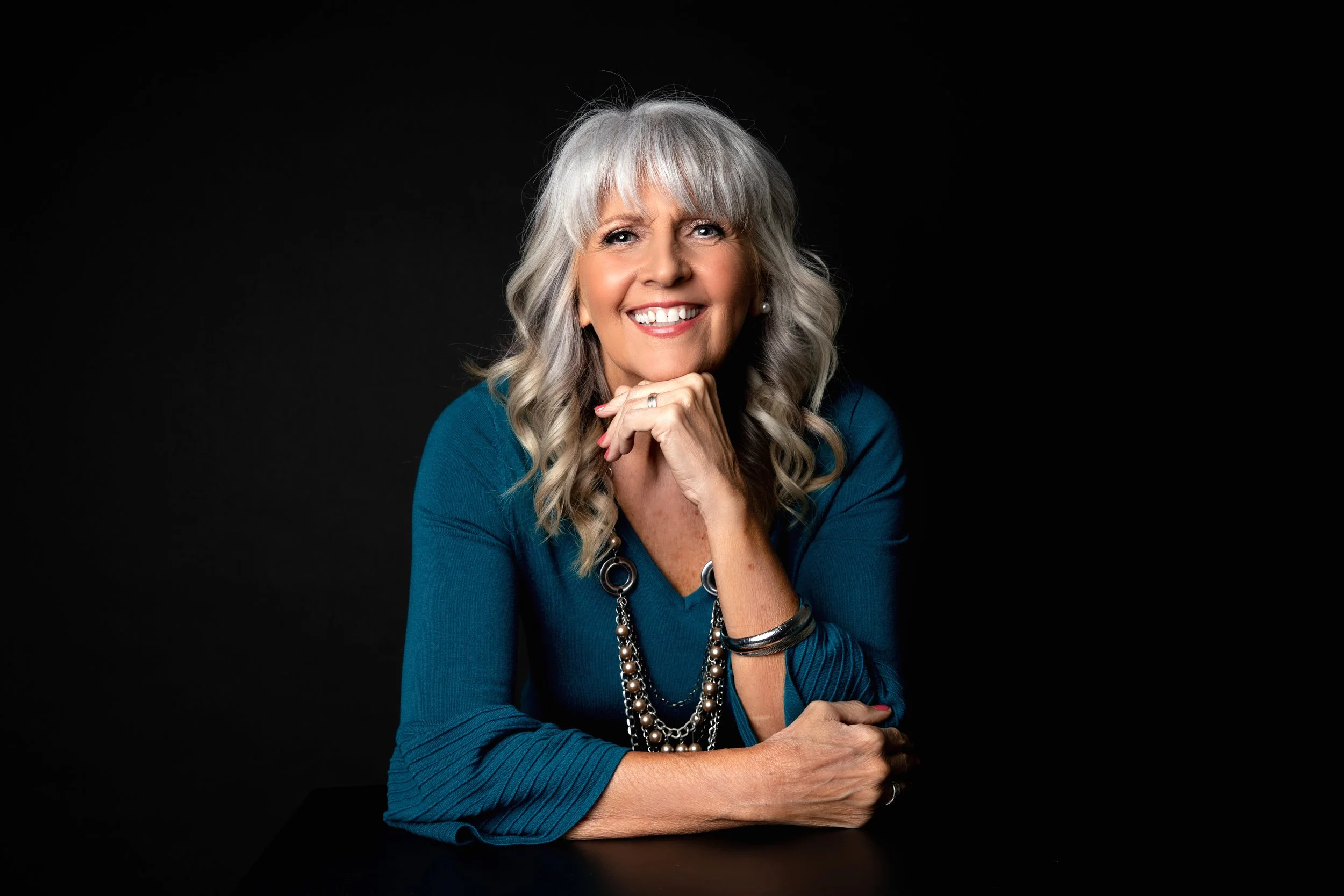 Portrait of an elegant woman with gray hair, wearing a blue blouse, jewelry, and smiling against a black background.