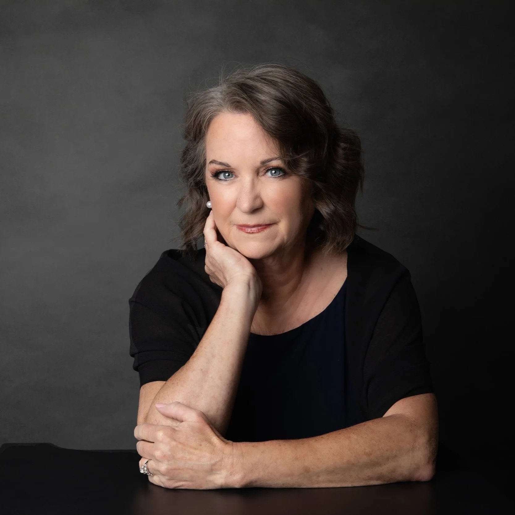 A middle-aged woman with wavy brown and gray hair posing with her chin resting on her hand against a dark gray background.