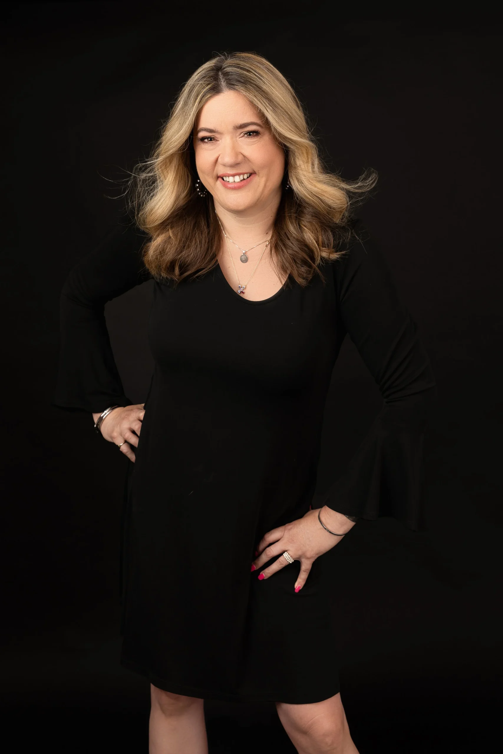 A woman with blonde and brown hair, smiling while standing against a black background, wearing a black dress and jewelry.