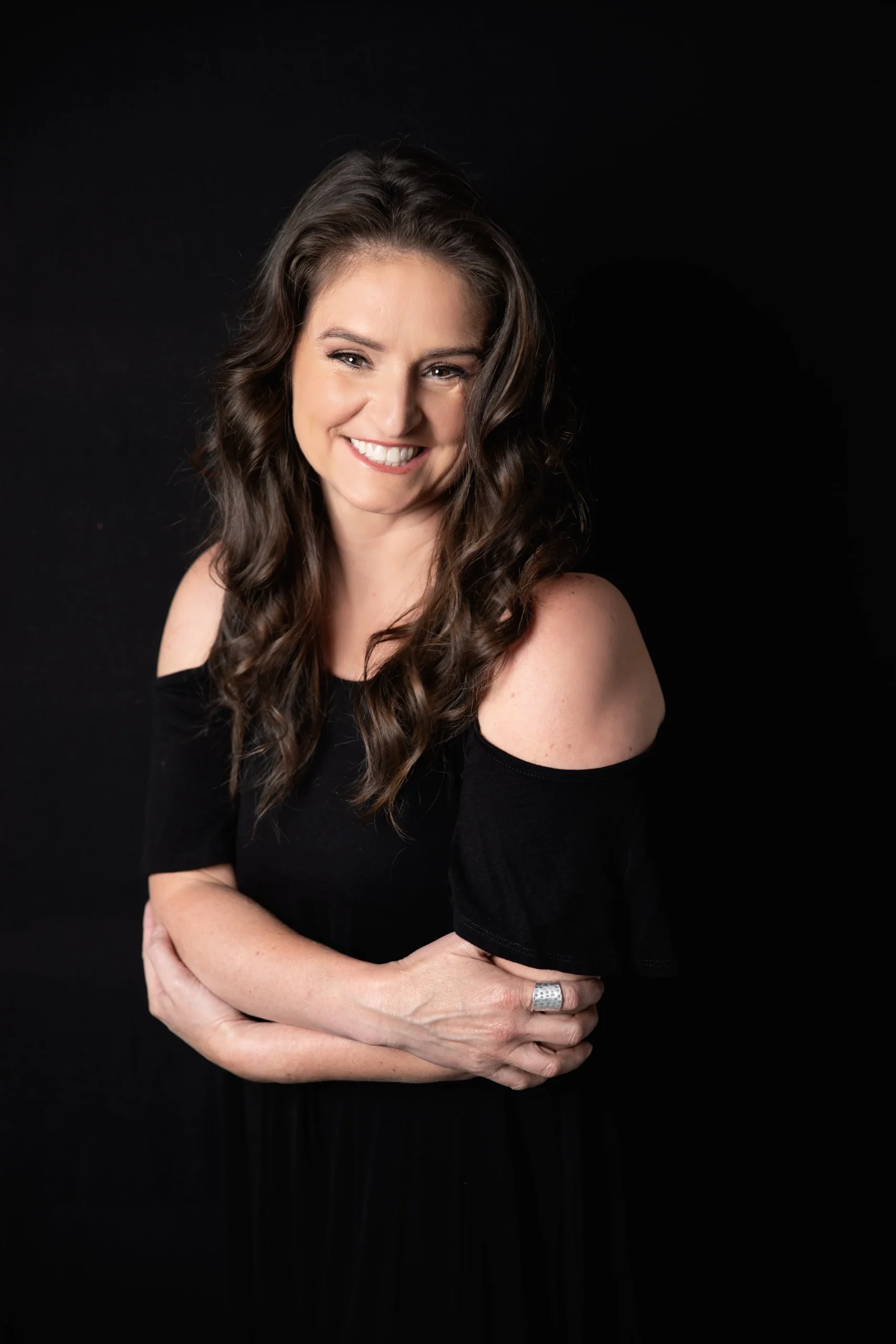 A woman with long, wavy brown hair, smiling, wearing a black off-the-shoulder dress, standing against a black background.