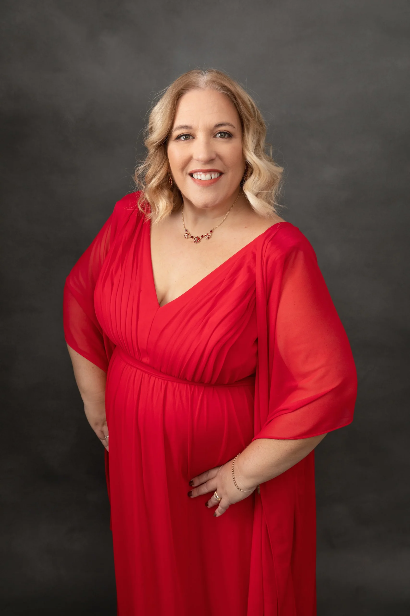 A woman with blonde, wavy hair wearing a red dress with sheer sleeves, smiling against a dark background.