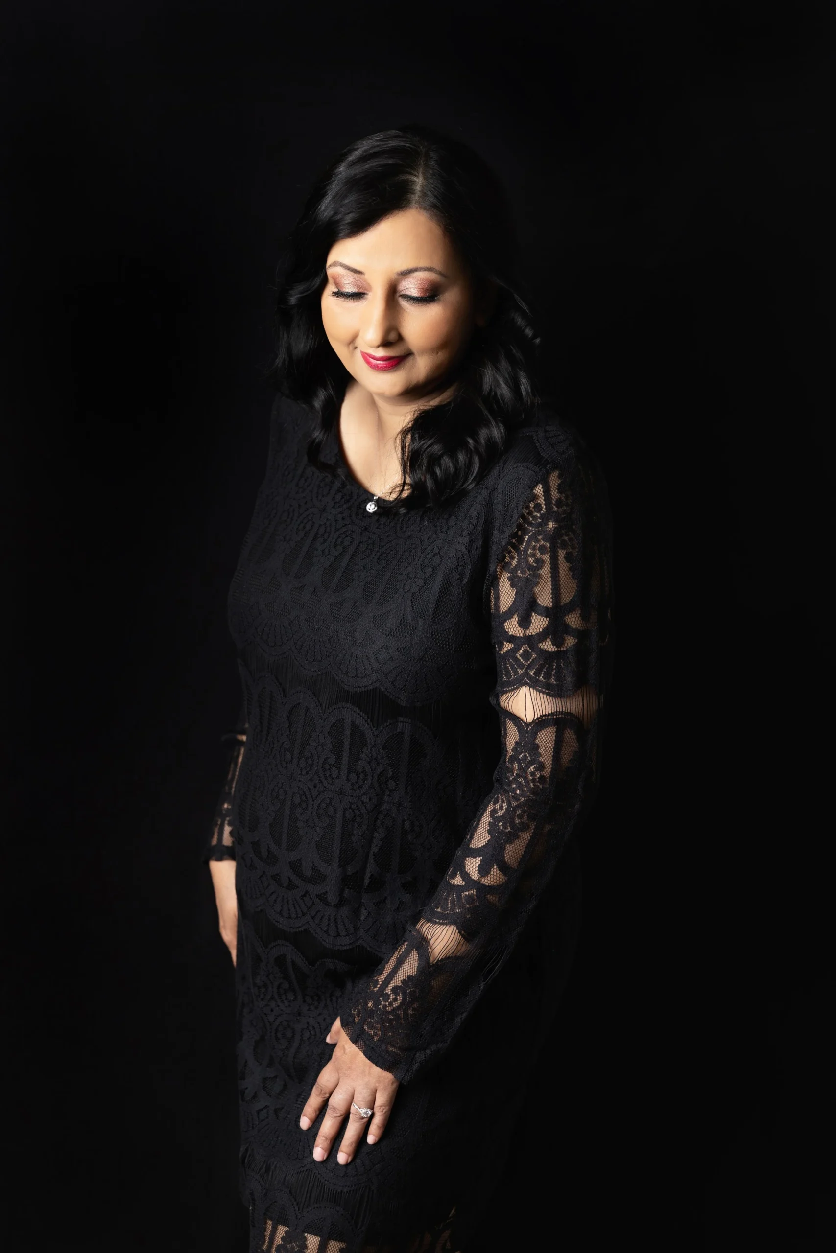 A woman with black hair and makeup, wearing a black lace dress, is looking down with her eyes closed against a black background.