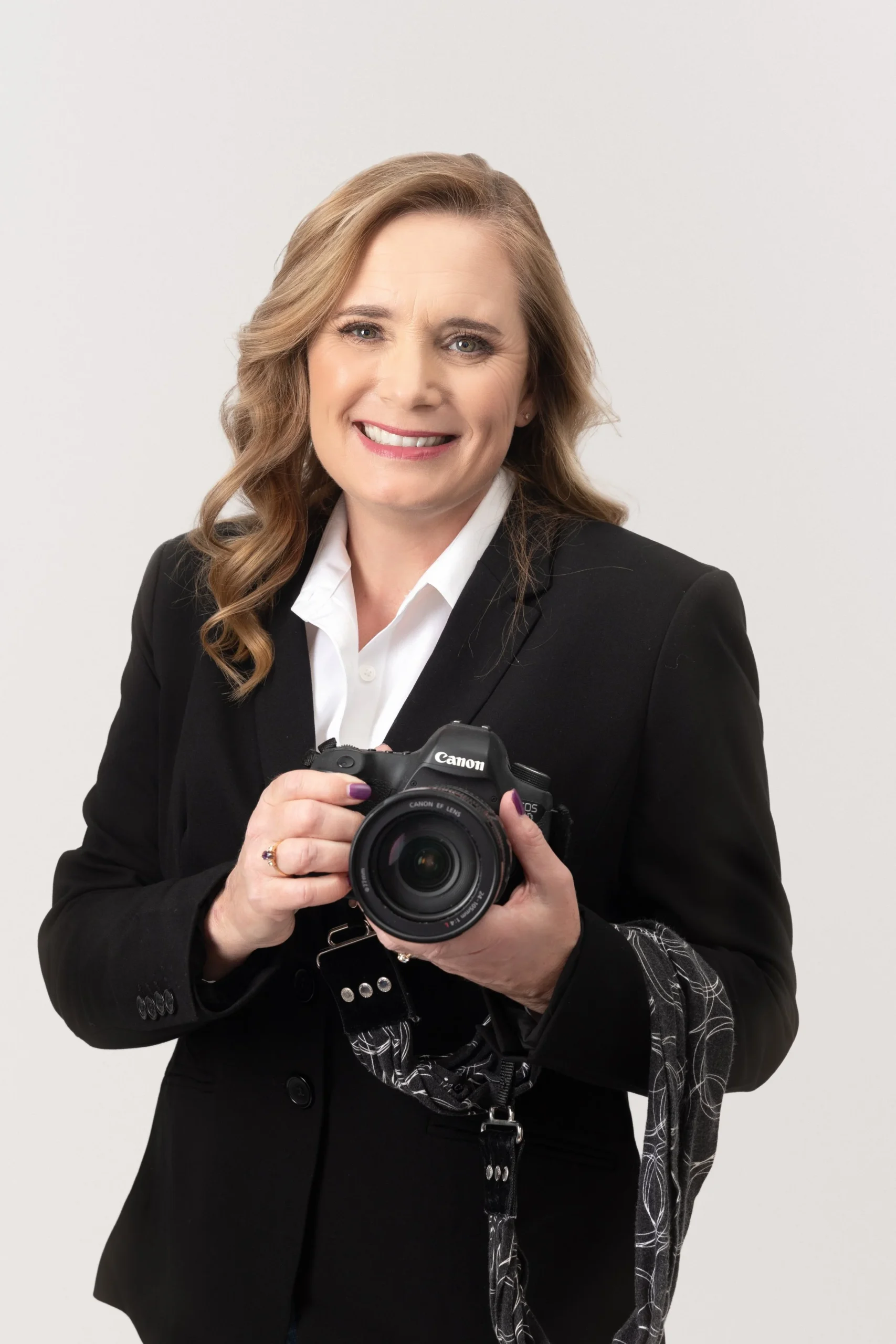 A woman with shoulder-length light brown hair, wearing a black blazer and white shirt, is holding a Canon camera and smiling at the camera against a plain light background.