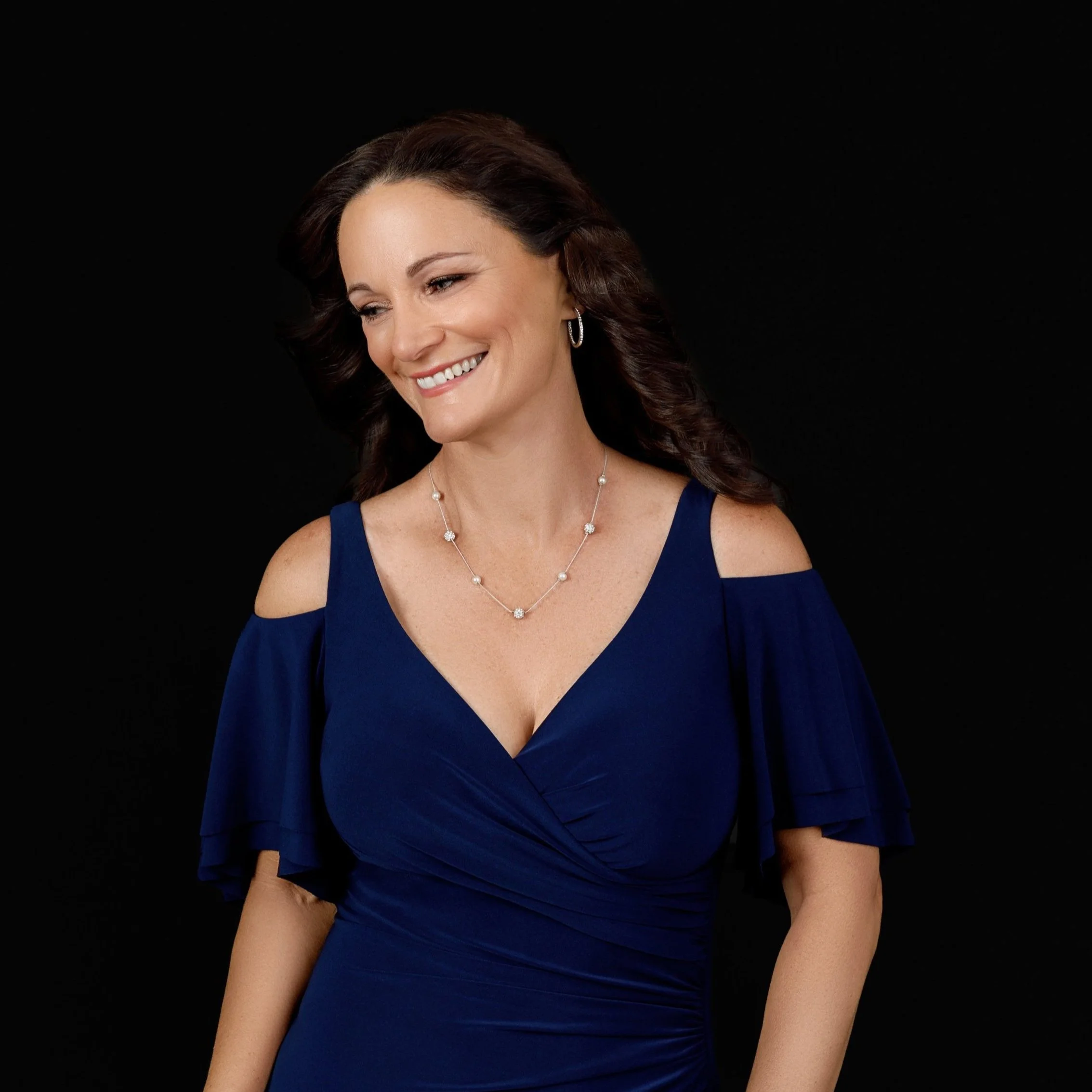 A woman with wavy brown hair, wearing a royal blue dress with open shoulders, smiling and looking down against a black background.