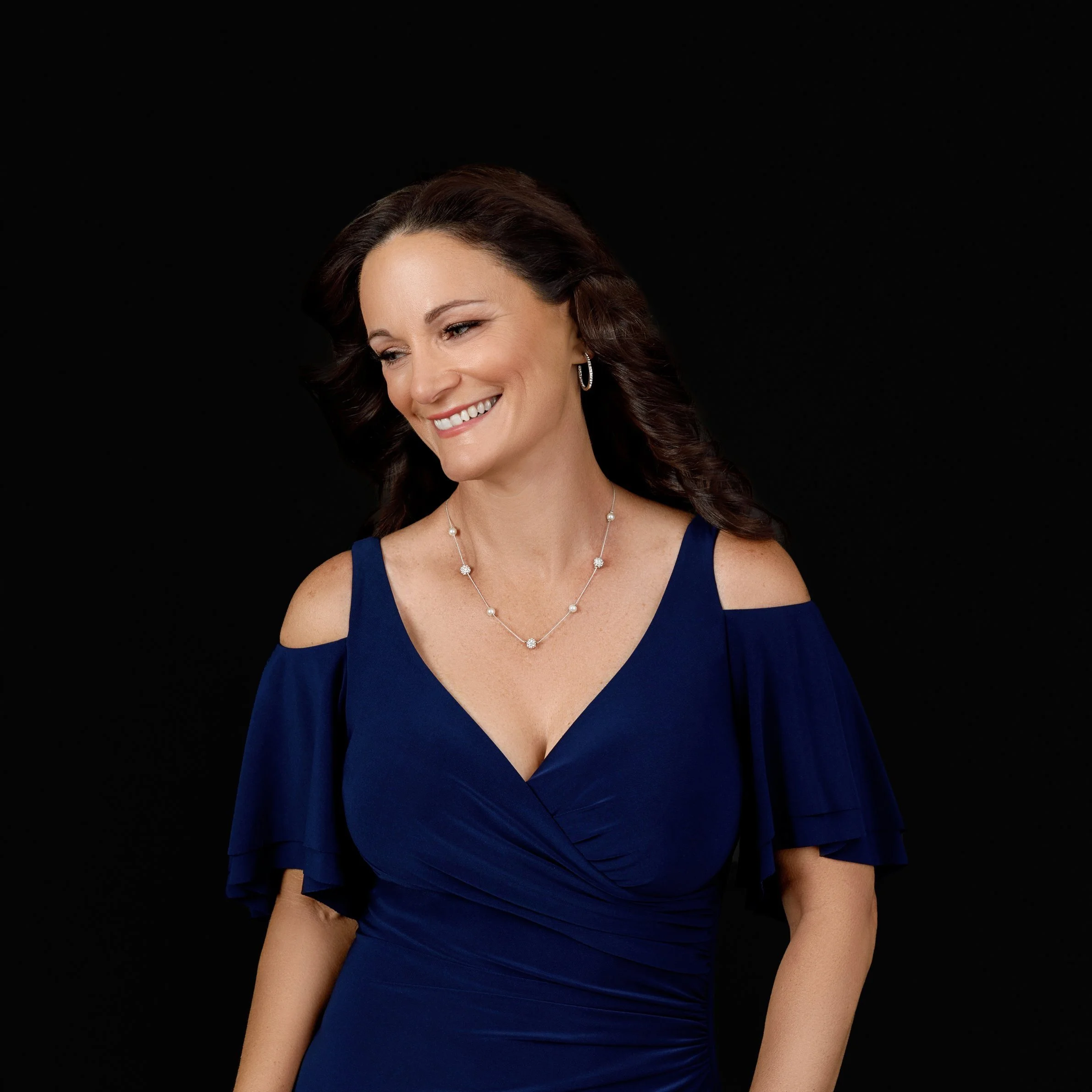 A woman smiling and looking to her left, wearing a dark blue dress with cut-out shoulders and jewelry, against a black background.