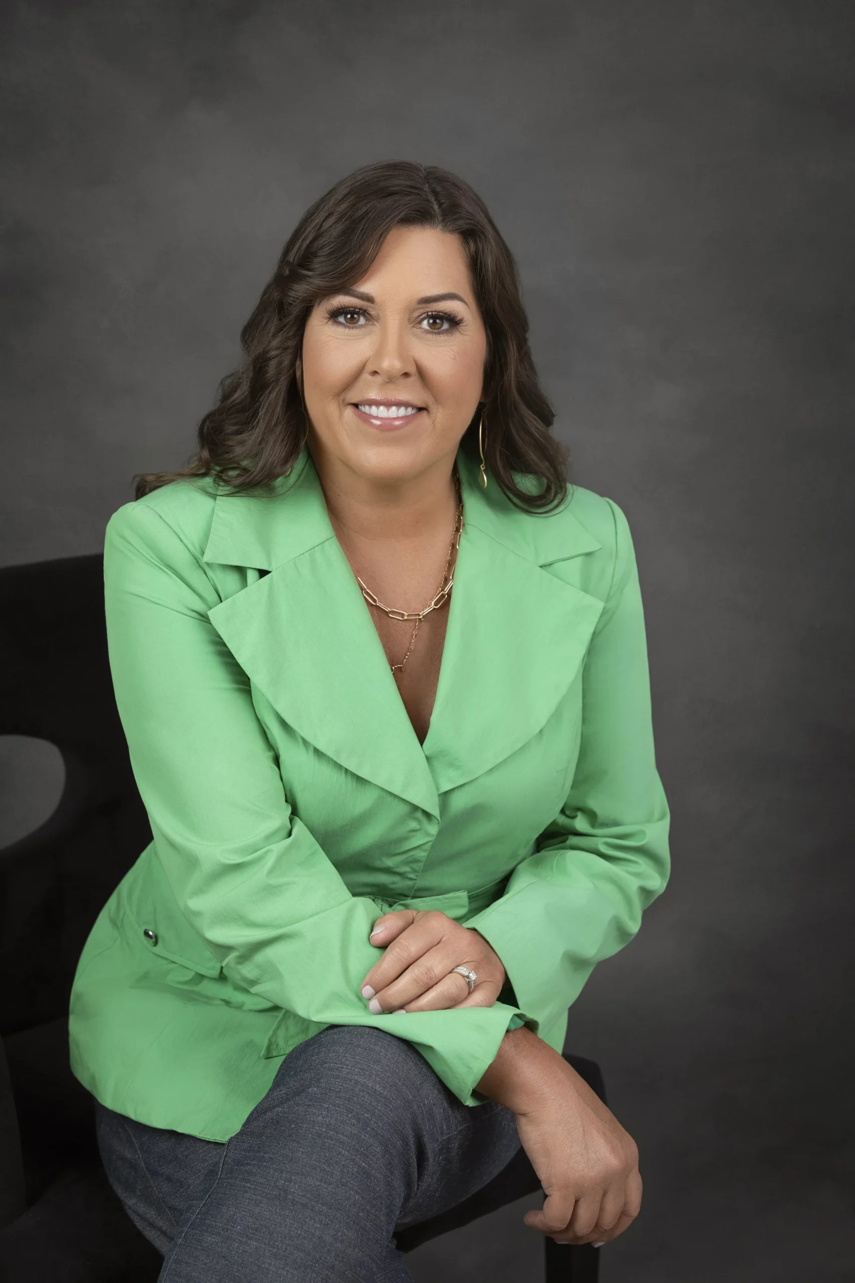 A woman with dark brown hair, wearing a bright green blazer and dark pants, sitting on a black chair, smiling at the camera against a gray background.