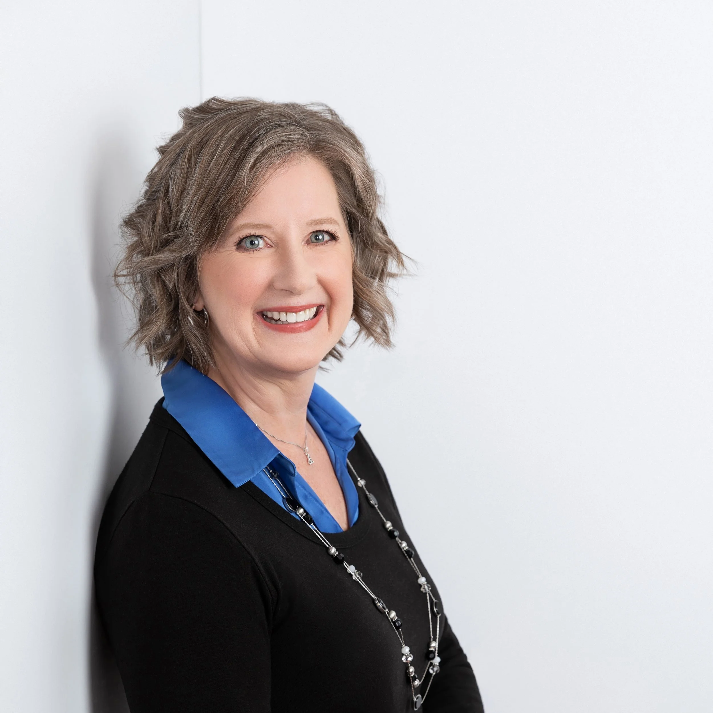 A smiling middle-aged woman with shoulder-length gray hair, wearing a black top over a blue collared shirt and a long necklace, standing against a plain white wall.