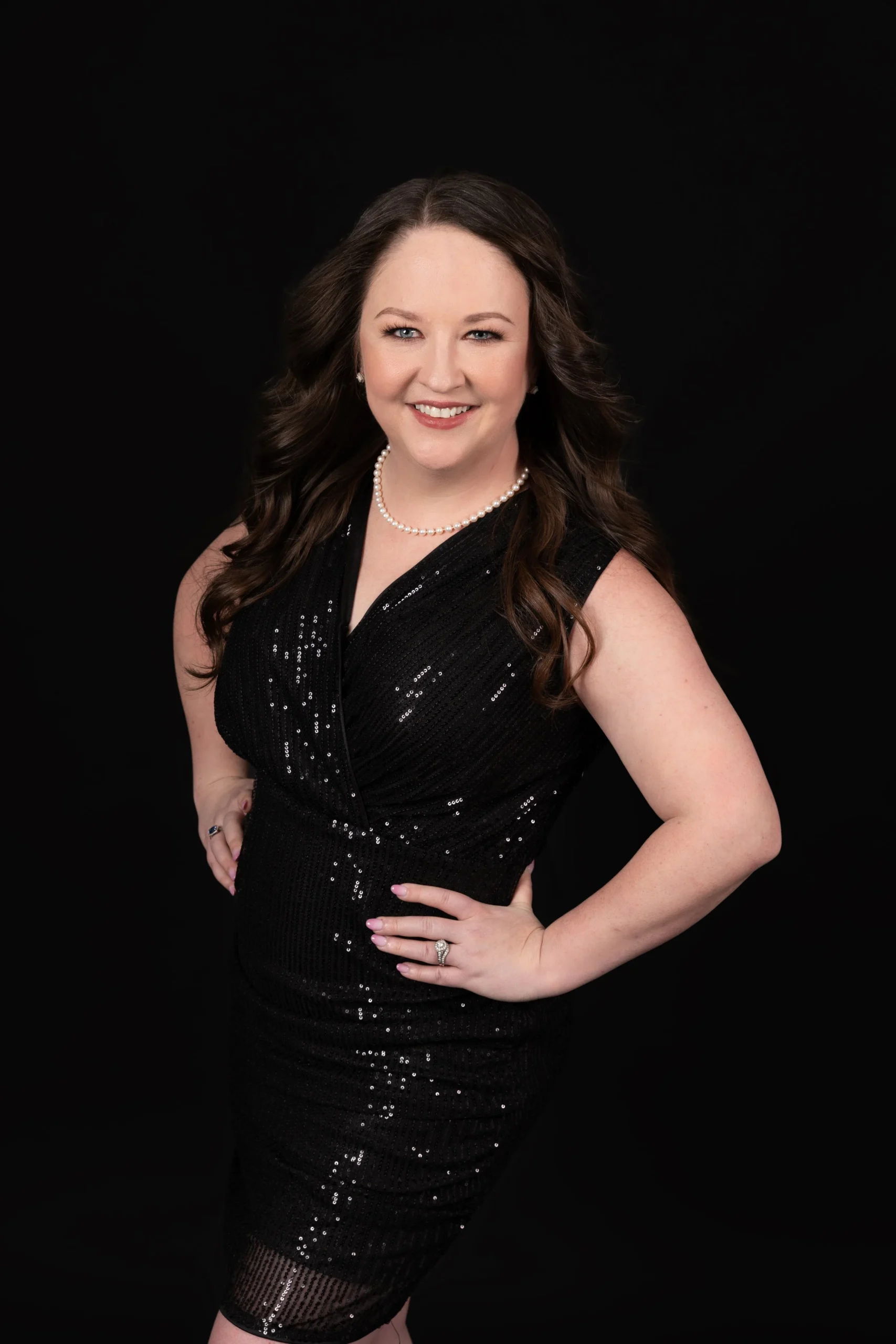 A woman with long brown hair, wearing a black, sleeveless, sequined dress, a pearl necklace, and a ring, smiling at the camera against a black background.