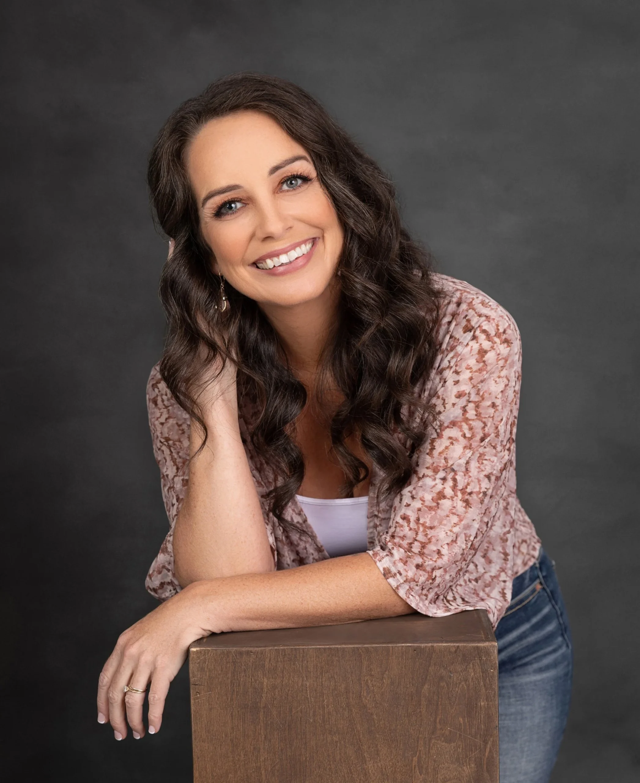 A woman with long curly brown hair and blue eyes smiling, resting her head on her left hand, leaning on a wooden surface, wearing a pink patterned top over a white shirt, with a dark gray background.