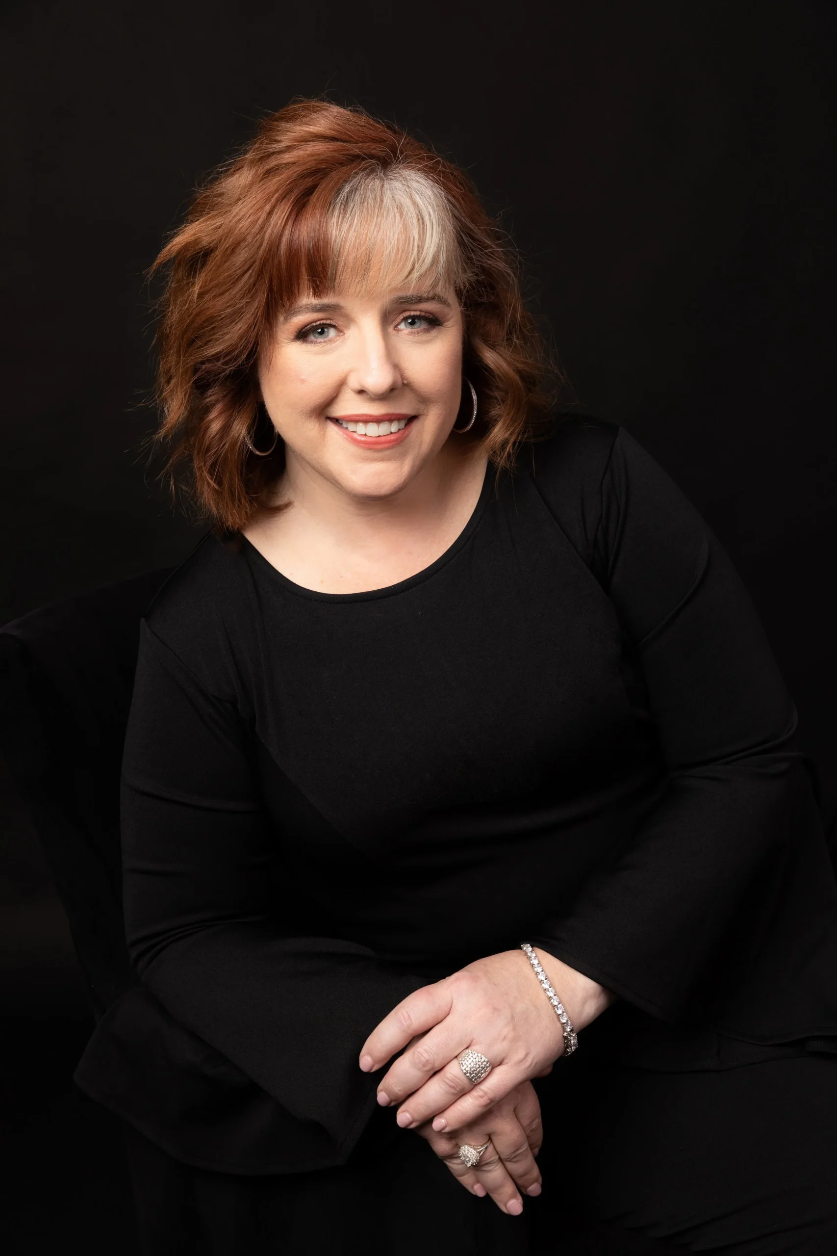 A woman with shoulder-length auburn hair, wearing a black long-sleeve top, sitting against a dark background, smiling, with jewelry including a bracelet, rings, and earrings.