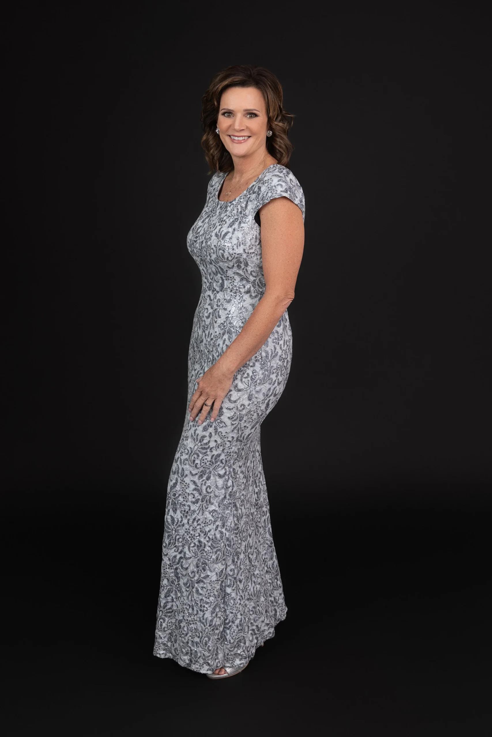 A woman in a silver, sequined, floor-length gown standing against a black background, smiling at the camera.