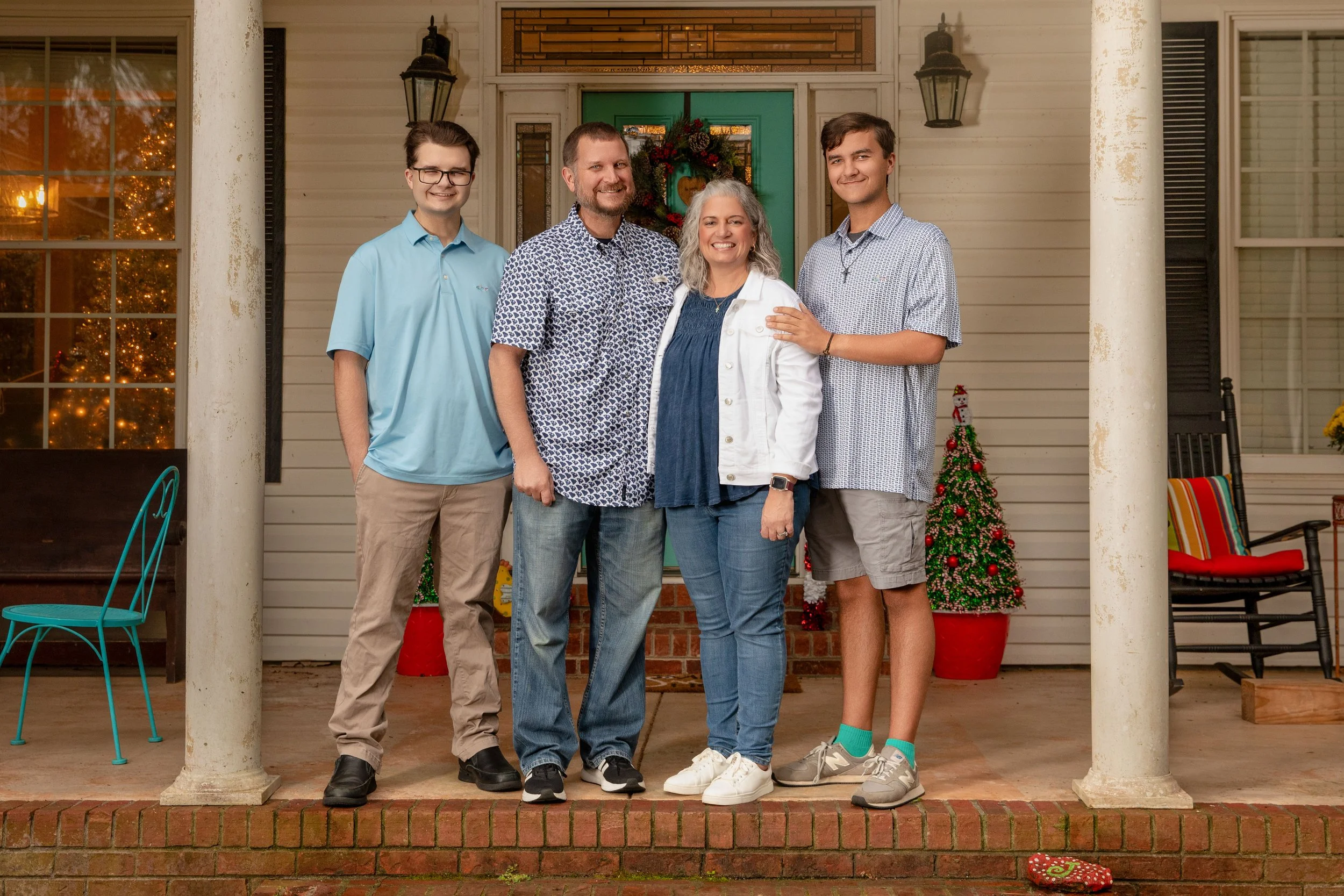 A family of five standing on a porch decorated for Christmas, with a Christmas tree, holiday wreath, and other festive decorations in the background.