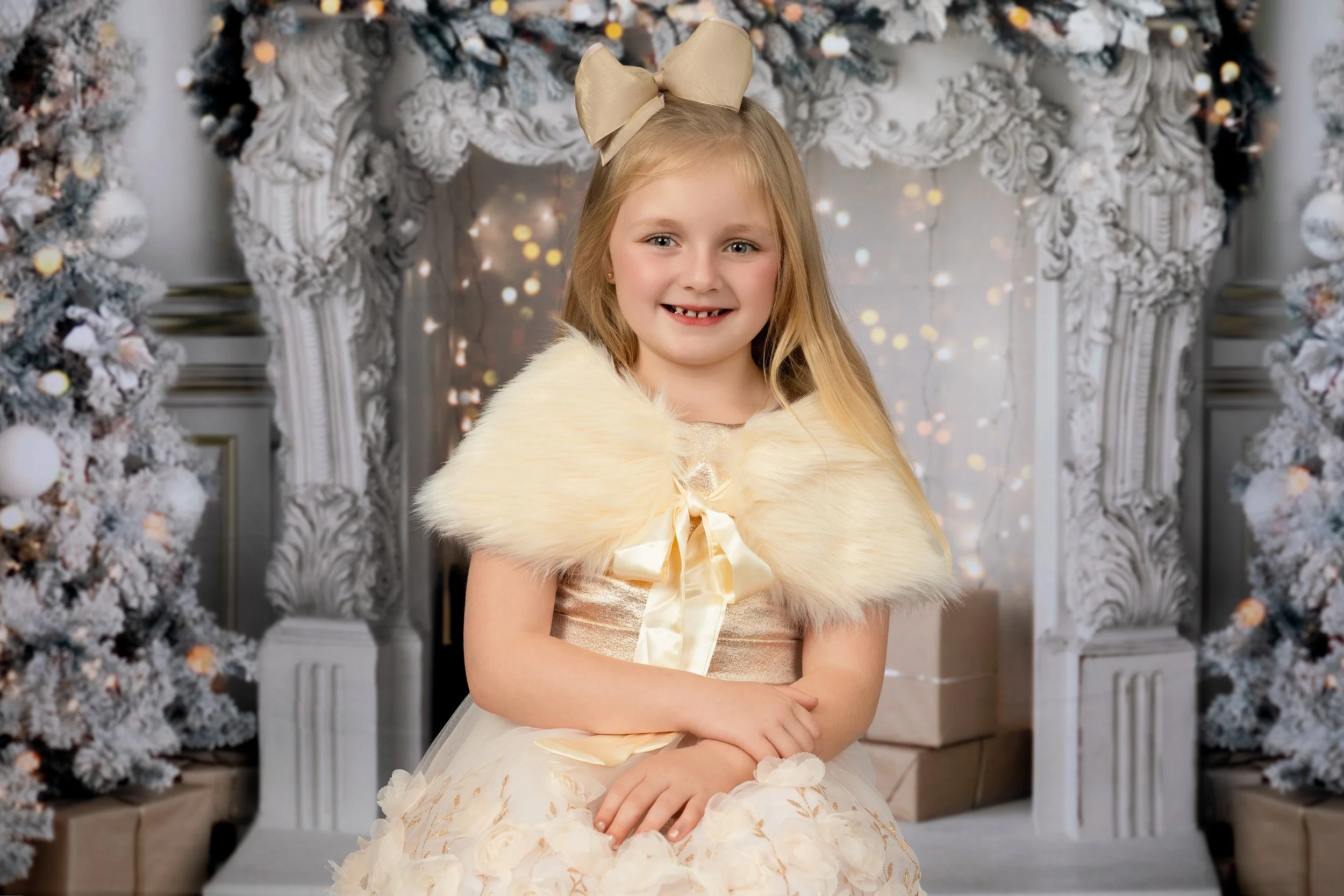 A young girl with long blonde hair, wearing a cream-colored dress with a satin ribbon and a faux fur shawl, sitting in front of a decorated white fireplace with Christmas trees and gifts, smiling at the camera.