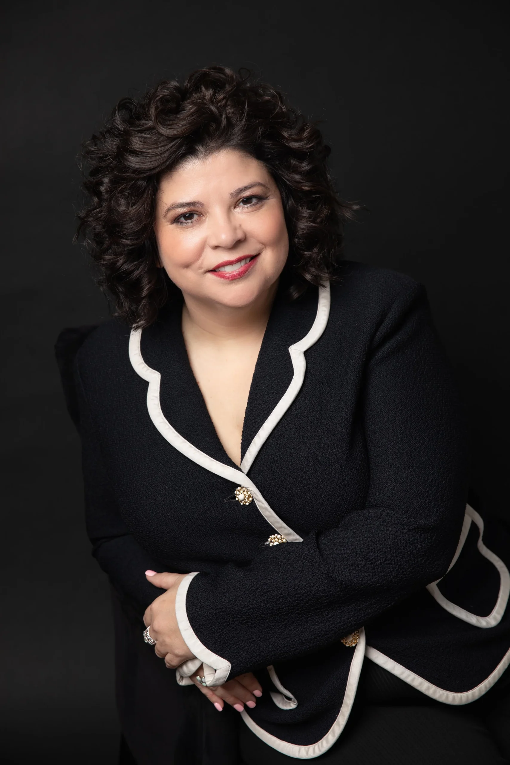 A woman with curly dark hair and light skin, smiling, wearing a black blazer with white piping and decorative buttons, against a black background.