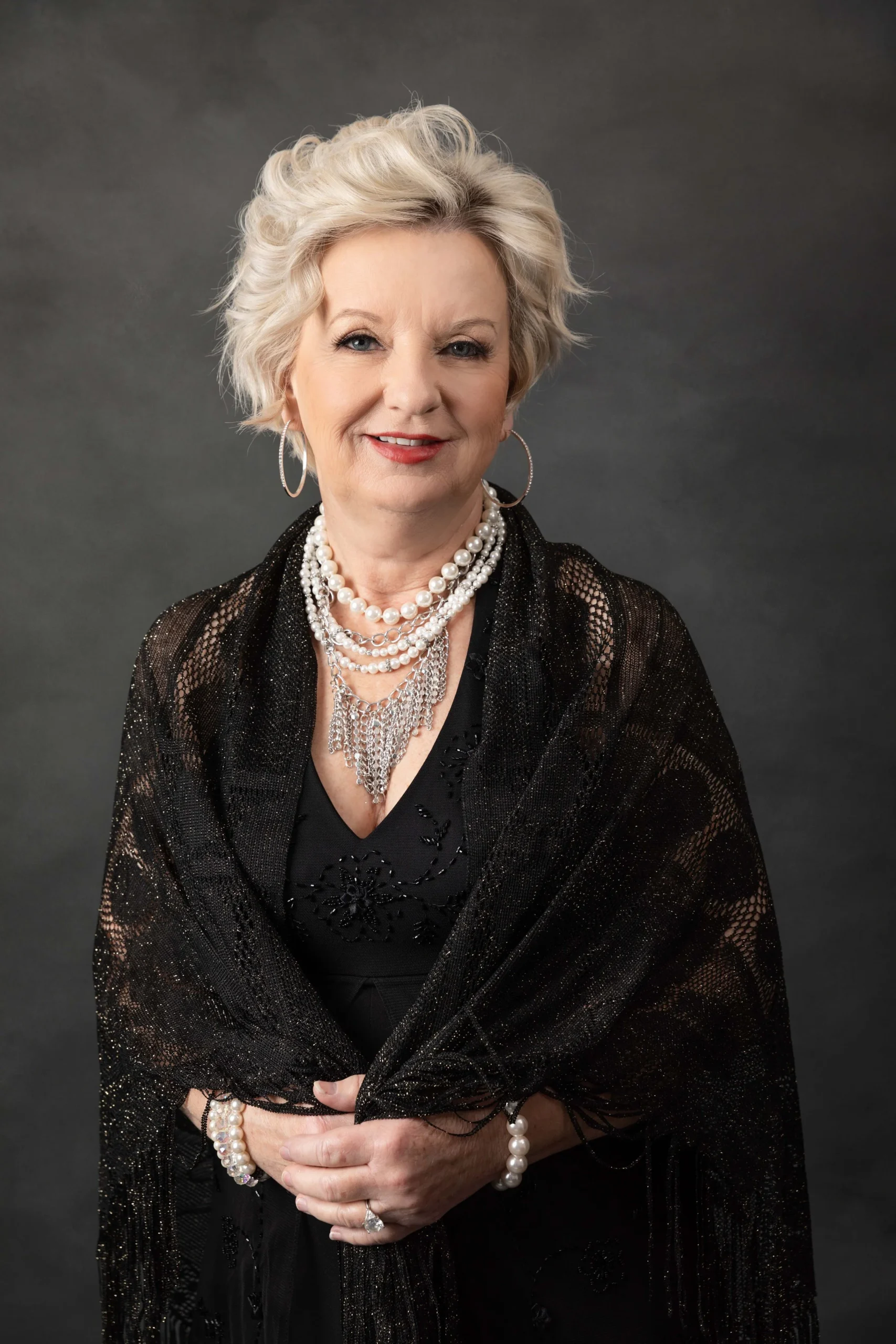 An elderly woman with short, curly, blonde hair smiling. She is dressed in a black outfit adorned with multiple pearl necklaces, earrings, bracelets, and a ring. She is wrapped in a black, glittery shawl, standing against a gray background.
