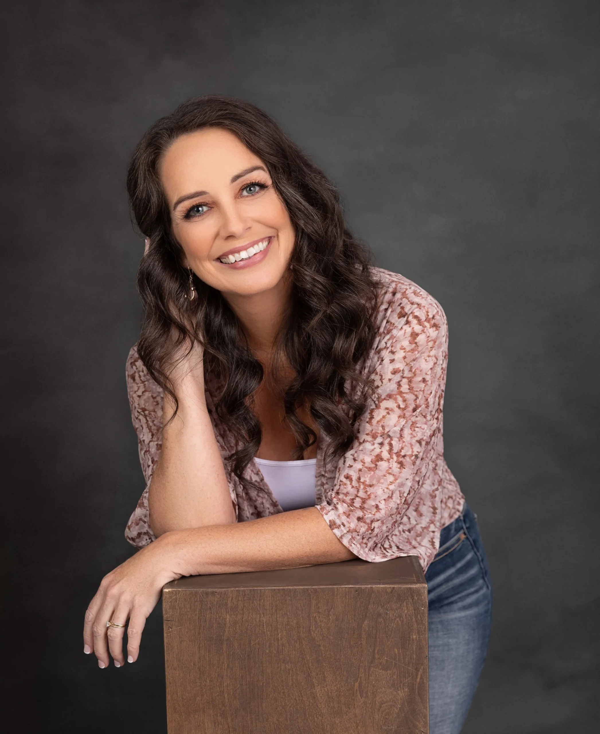 A woman with long, dark curly hair, smiling, wearing a light pink patterned blouse and blue jeans, leaning on a wooden block against a gray background.