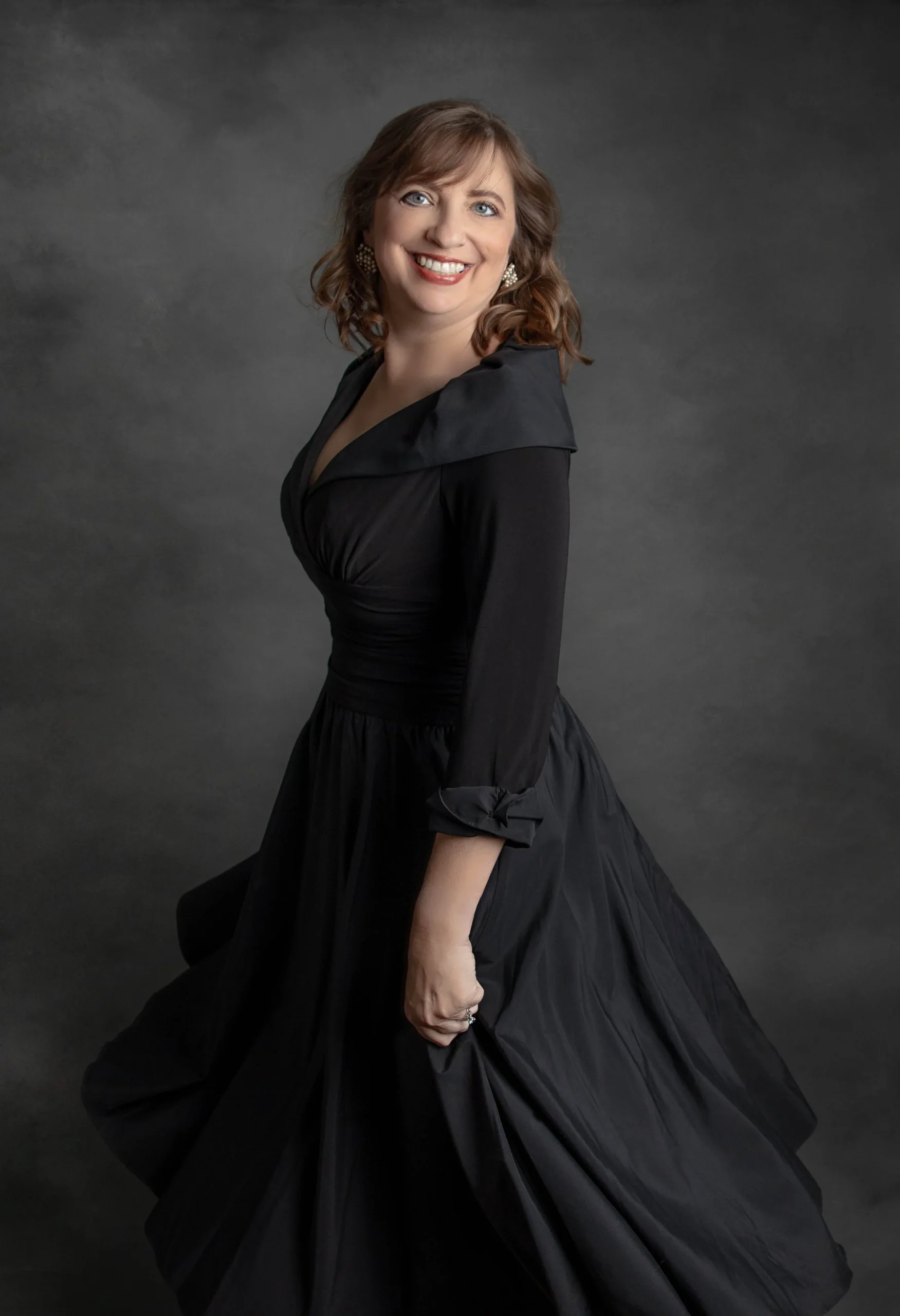 A woman in a black dress standing against a gray background, smiling, with her body slightly turned and her right arm bent.