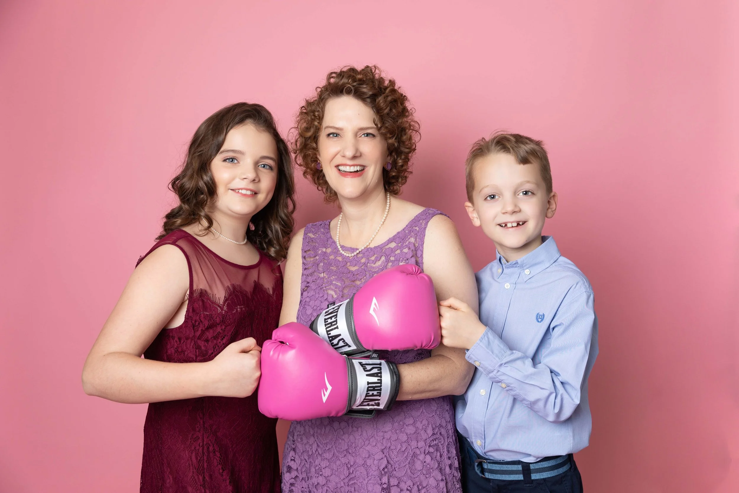A woman wearing pink boxing gloves stands between a girl in a burgundy dress and a boy in a blue shirt, all smiling against a pink background.