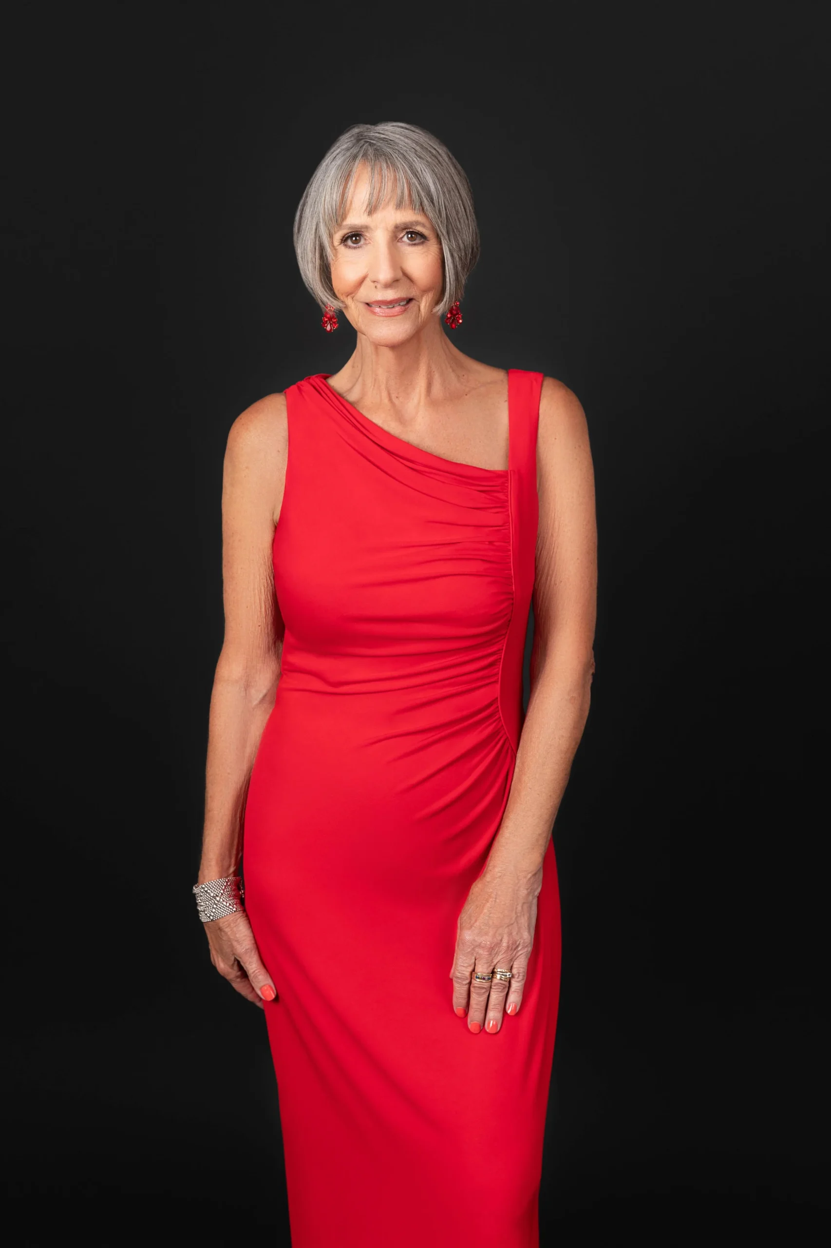An elderly woman with short gray hair in a red sleeveless dress with jewelry against a black background.