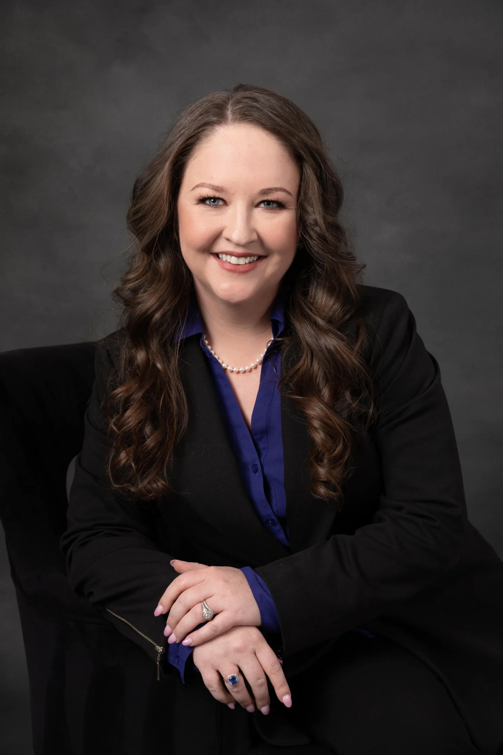 Portrait of a woman with long, wavy brown hair wearing a black blazer, blue blouse, pearl necklace, and rings, smiling against a dark gray background.