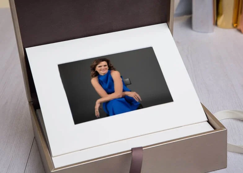 A framed photograph of a woman in a blue dress sitting and smiling, placed inside a cardboard box with a white border and a ribbon, on a light-colored floor.