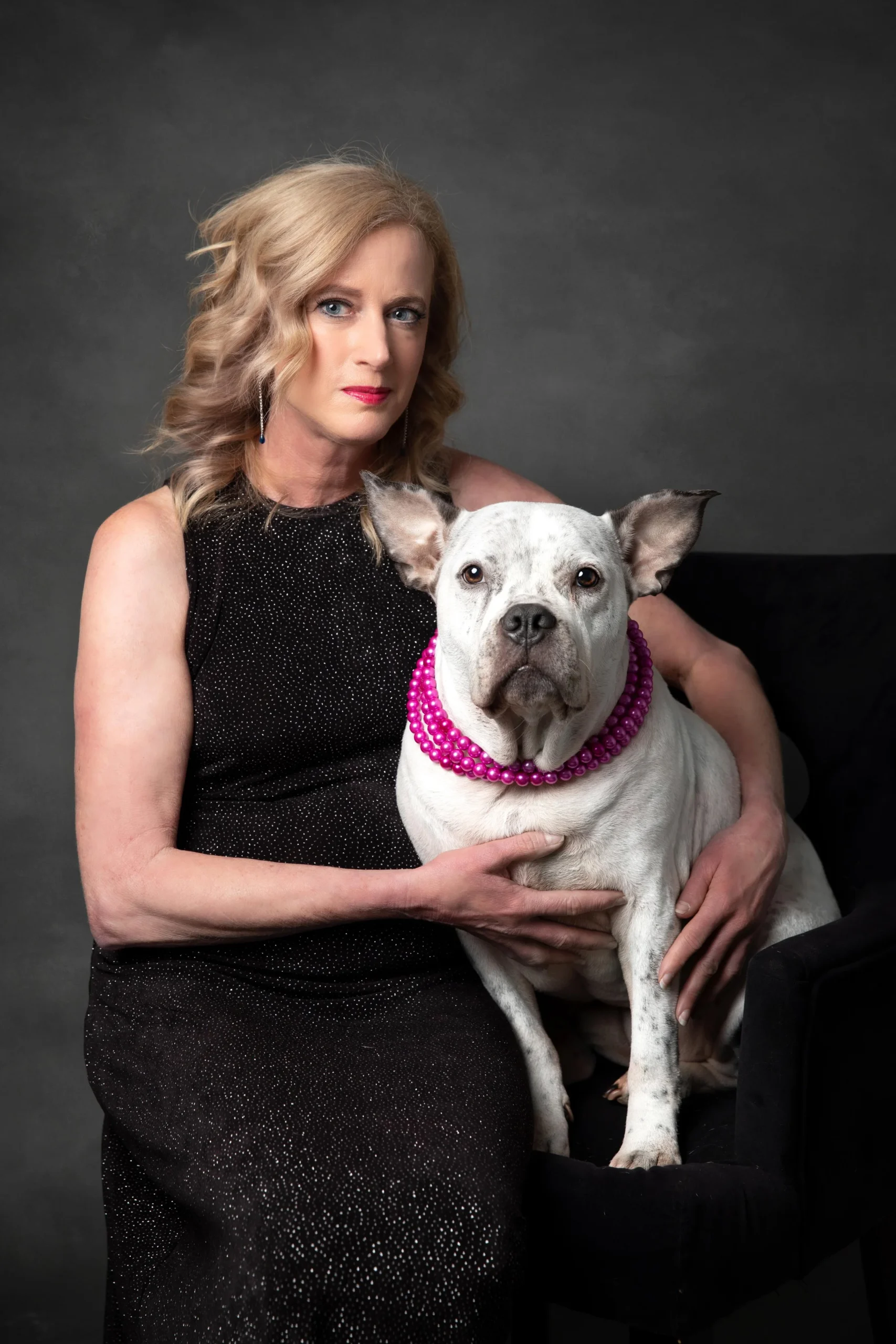 A woman with blonde hair wearing a black, sparkly sleeveless dress is holding a white dog with gray spots and large ears that is wearing a pink beaded collar. The background is dark gray.