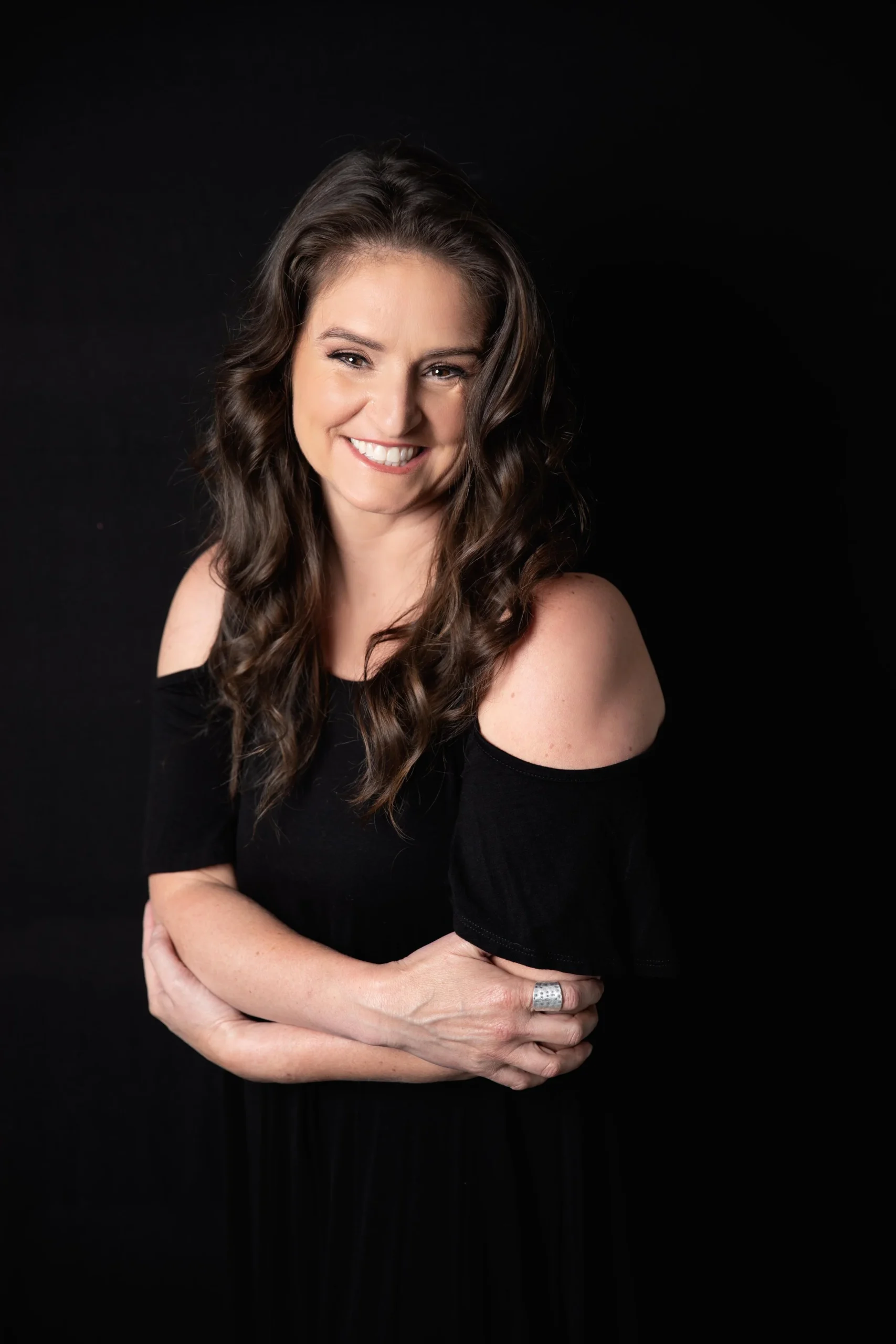 A woman with long, wavy brown hair, smiling and wearing a black off-shoulder dress, standing against a black background.