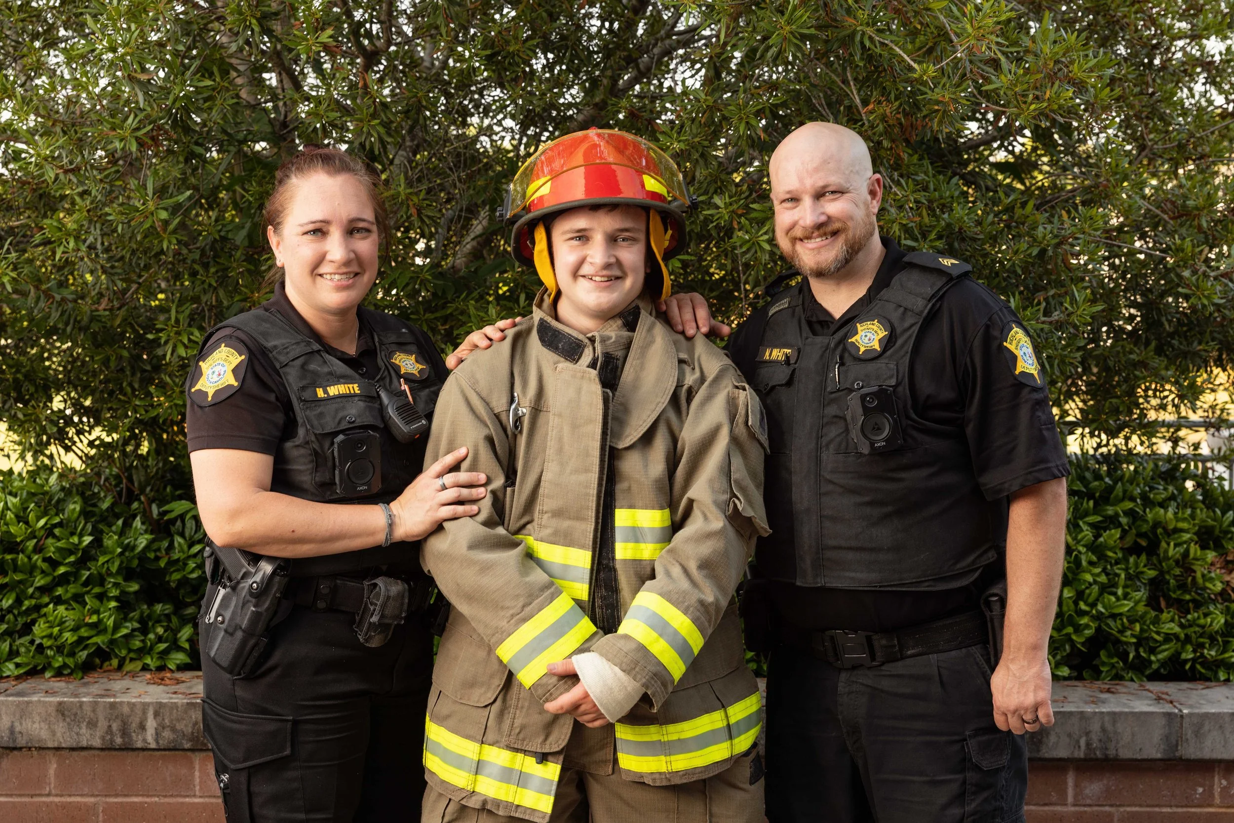 A firefighter in full gear with two police officers, one woman and one man, standing outdoors in front of green bushes on a sunny day.