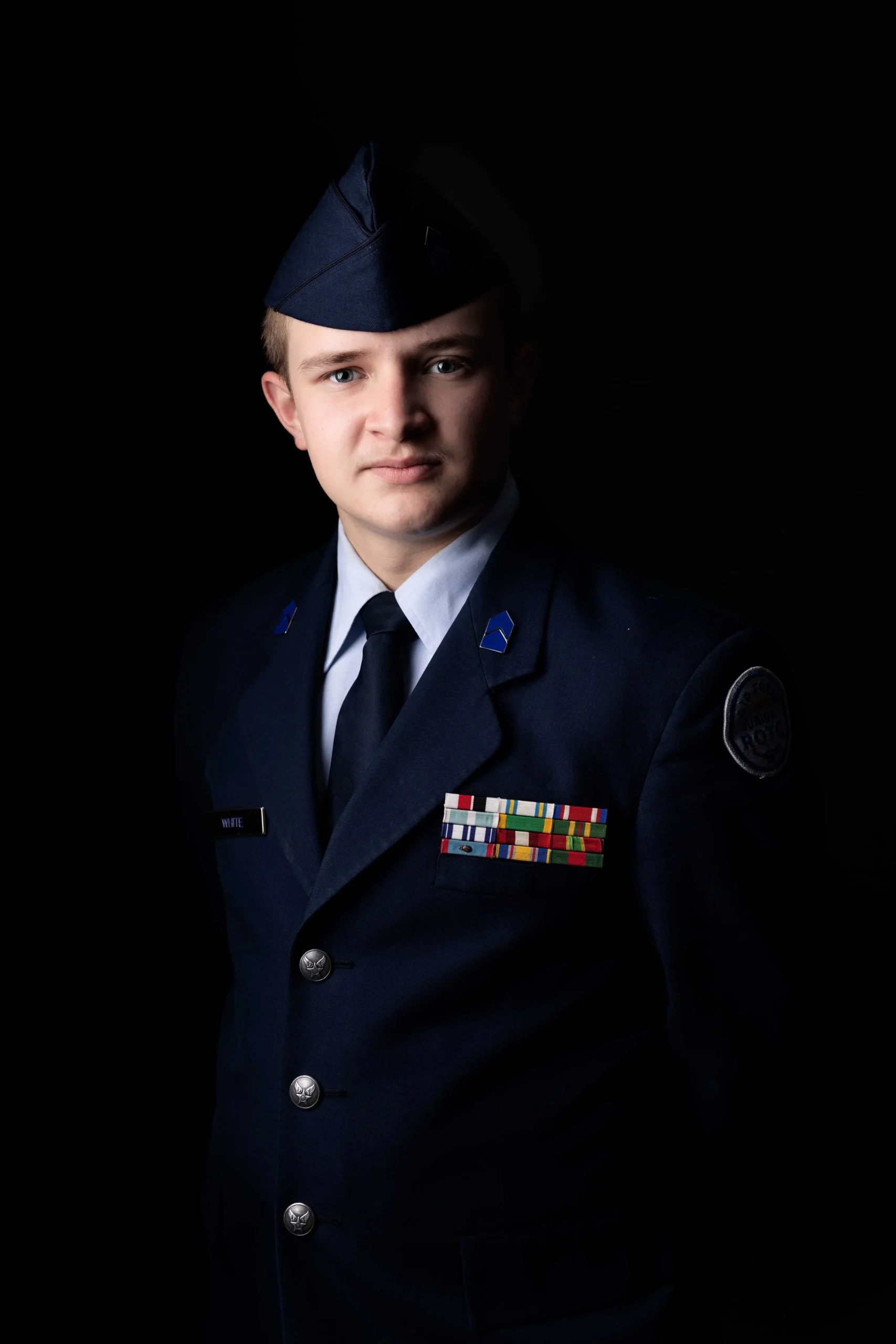 Young male police officer in uniform, with military-style hat, dark jacket, and colorful ribbons on chest, against a dark background.