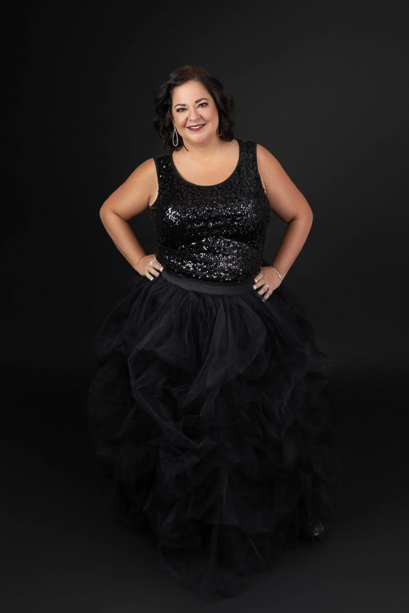 A woman in a black glittery sleeveless top and a voluminous black tulle skirt, smiling and posing with hands on her hips against a plain black background.