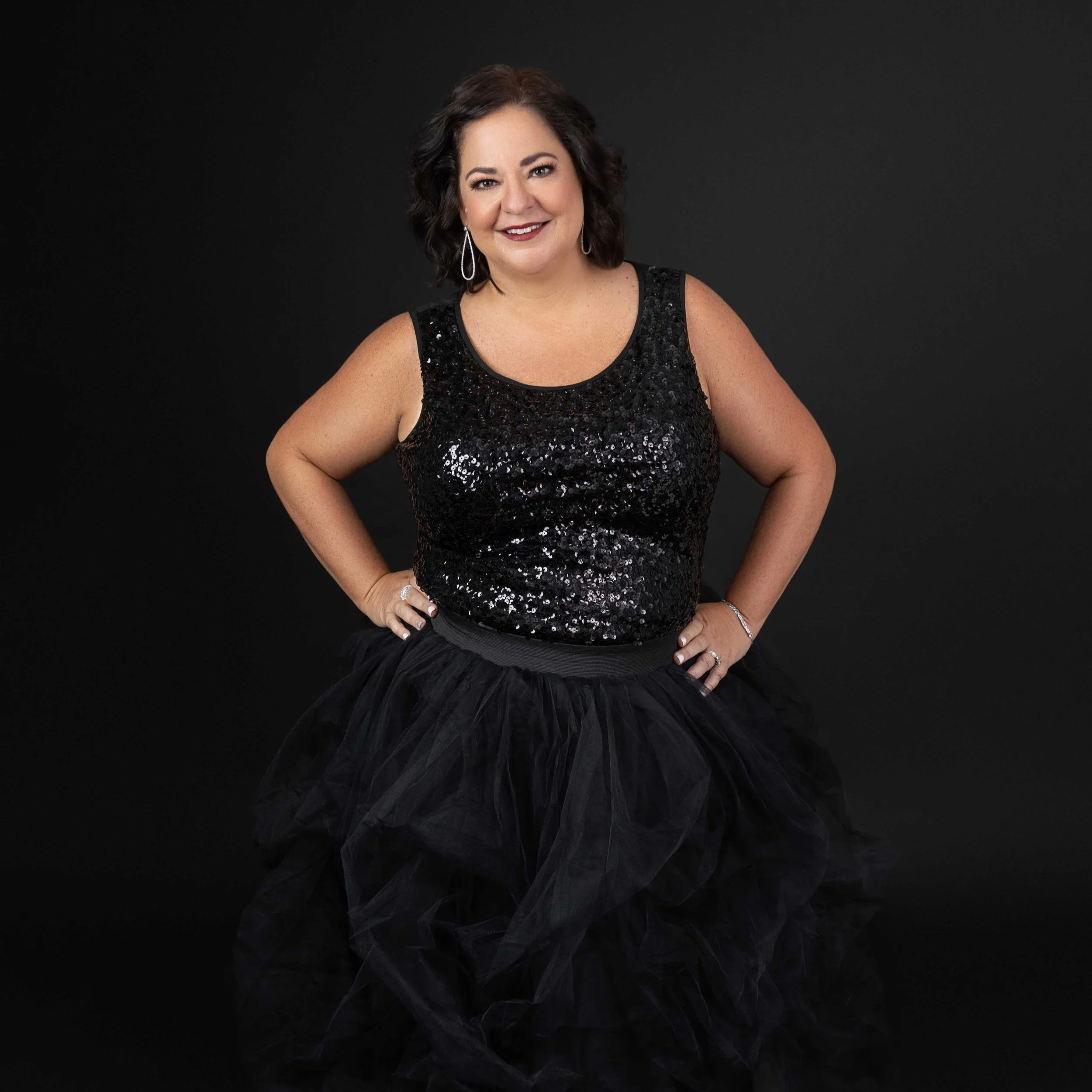 A woman with dark hair in loose curls, wearing a sleeveless black sequin top and a black tulle skirt, smiles with hands on hips against a black background.