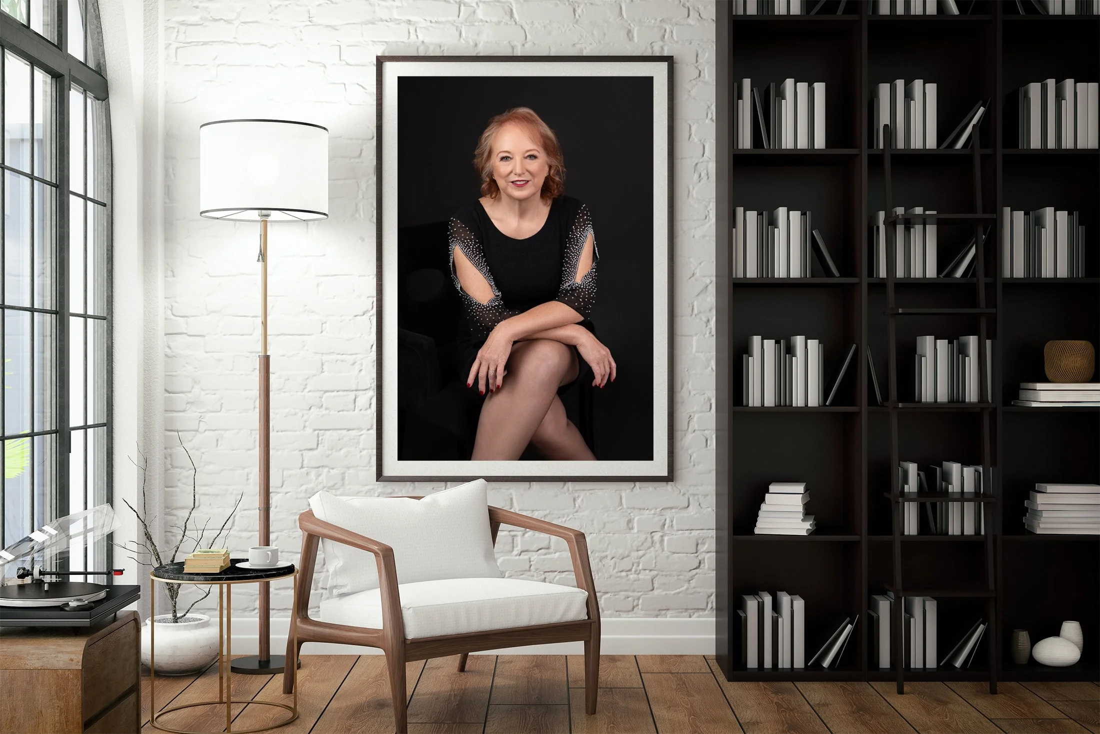 Living room with a large framed portrait of a woman on a white brick wall, a tall floor lamp, a white armchair, a small side table with a coffee mug and books, a turntable on a wooden stand, and a black bookshelf filled with books and decorative items.
