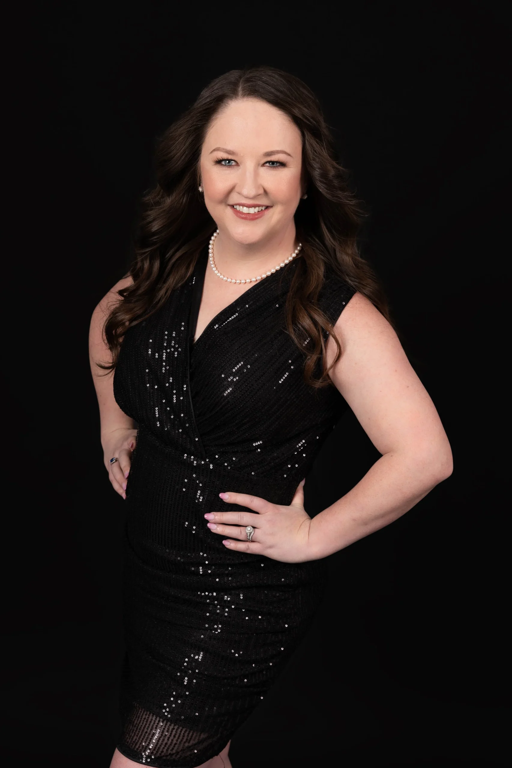 A woman with long wavy brown hair, wearing a black sequin dress, pearl necklace, and rings, posing against a black background.
