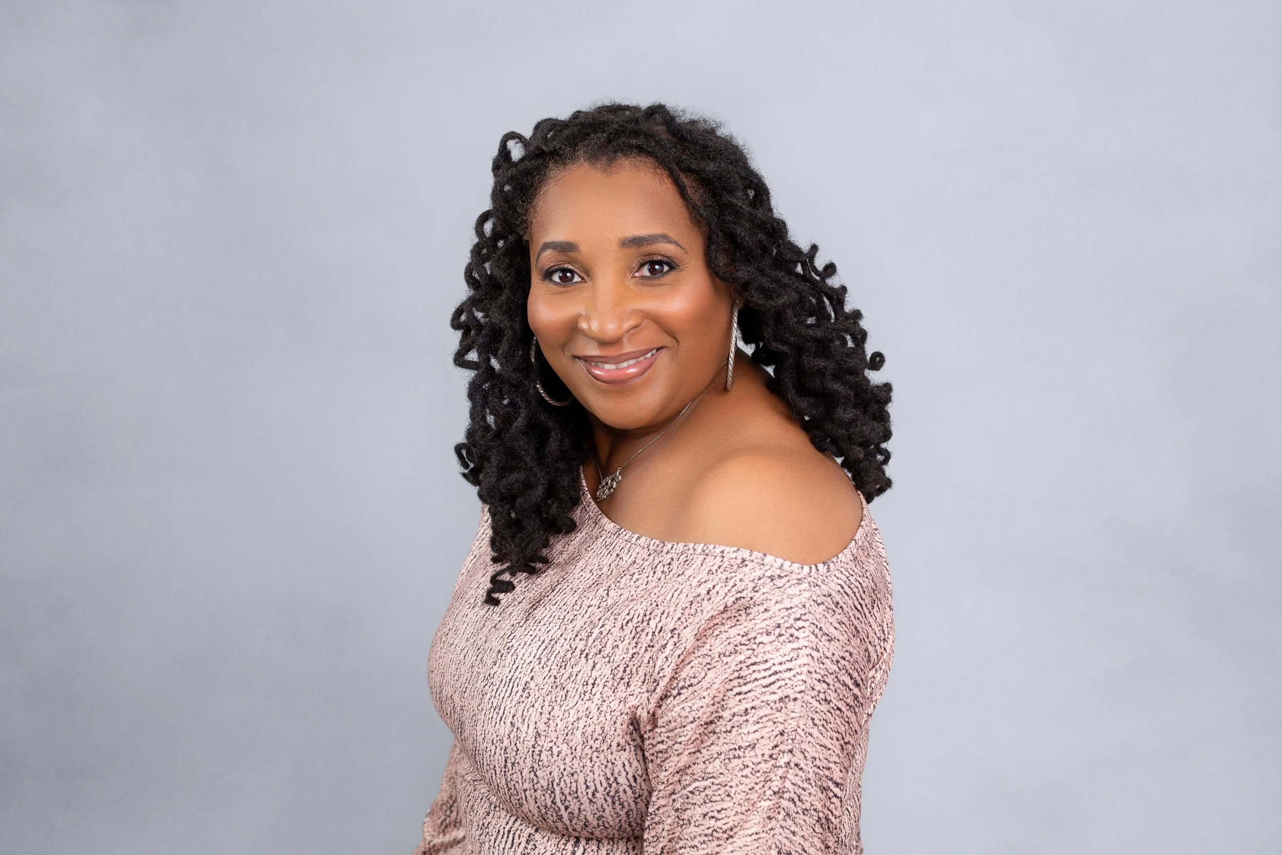 A woman with curly black hair smiling and wearing a pink off-the-shoulder top standing against a plain light gray background.