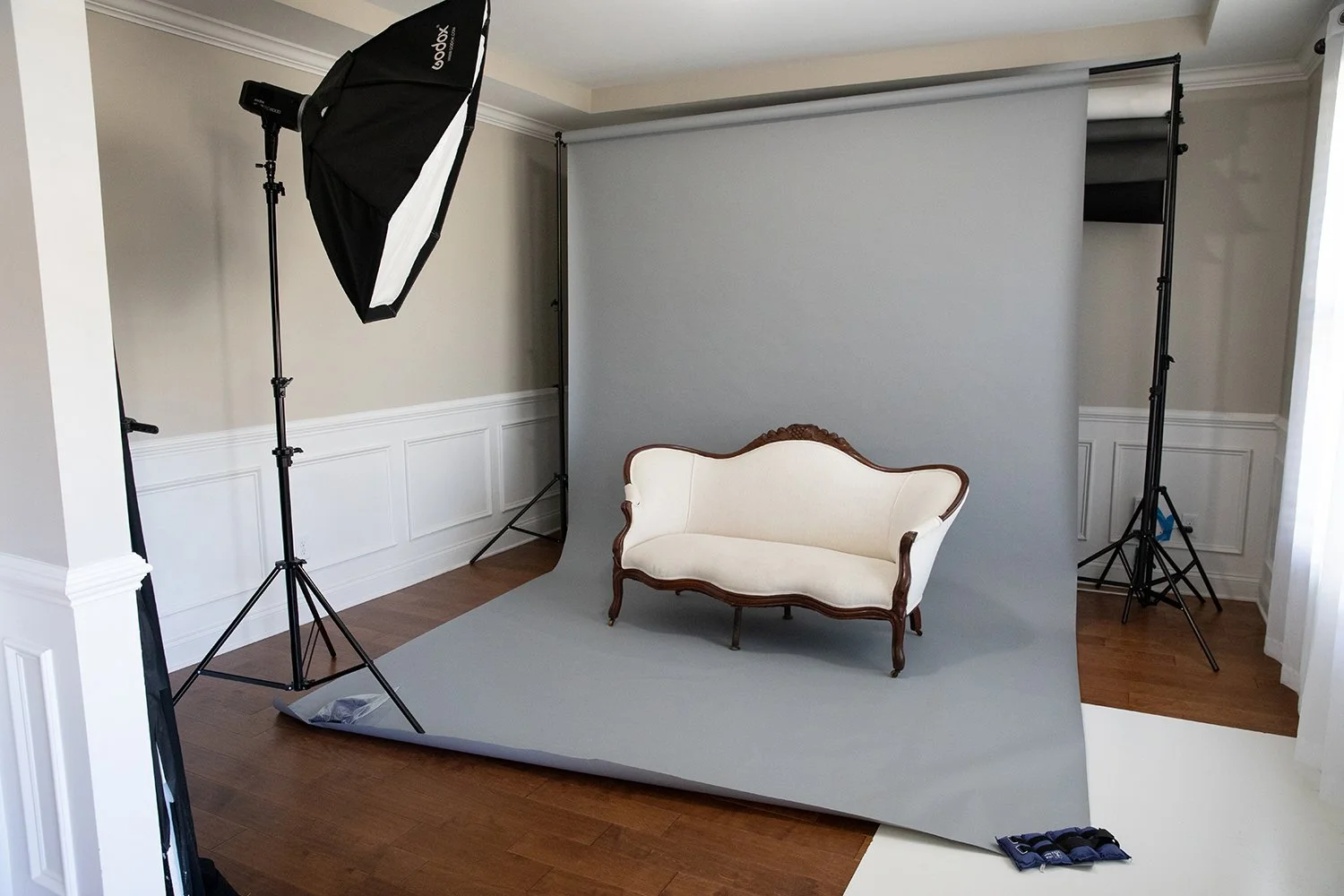 Photography studio with a grey backdrop, professional lighting equipment, and a vintage white sofa with wooden trim.