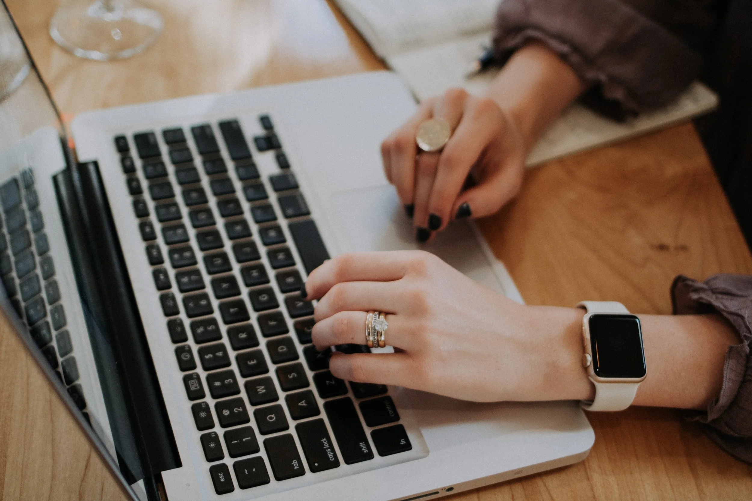 A person typing on a silver laptop with a white band smartwatch on their wrist, a large ring on their finger, and a gold ring on their middle finger, sitting at a wooden table.